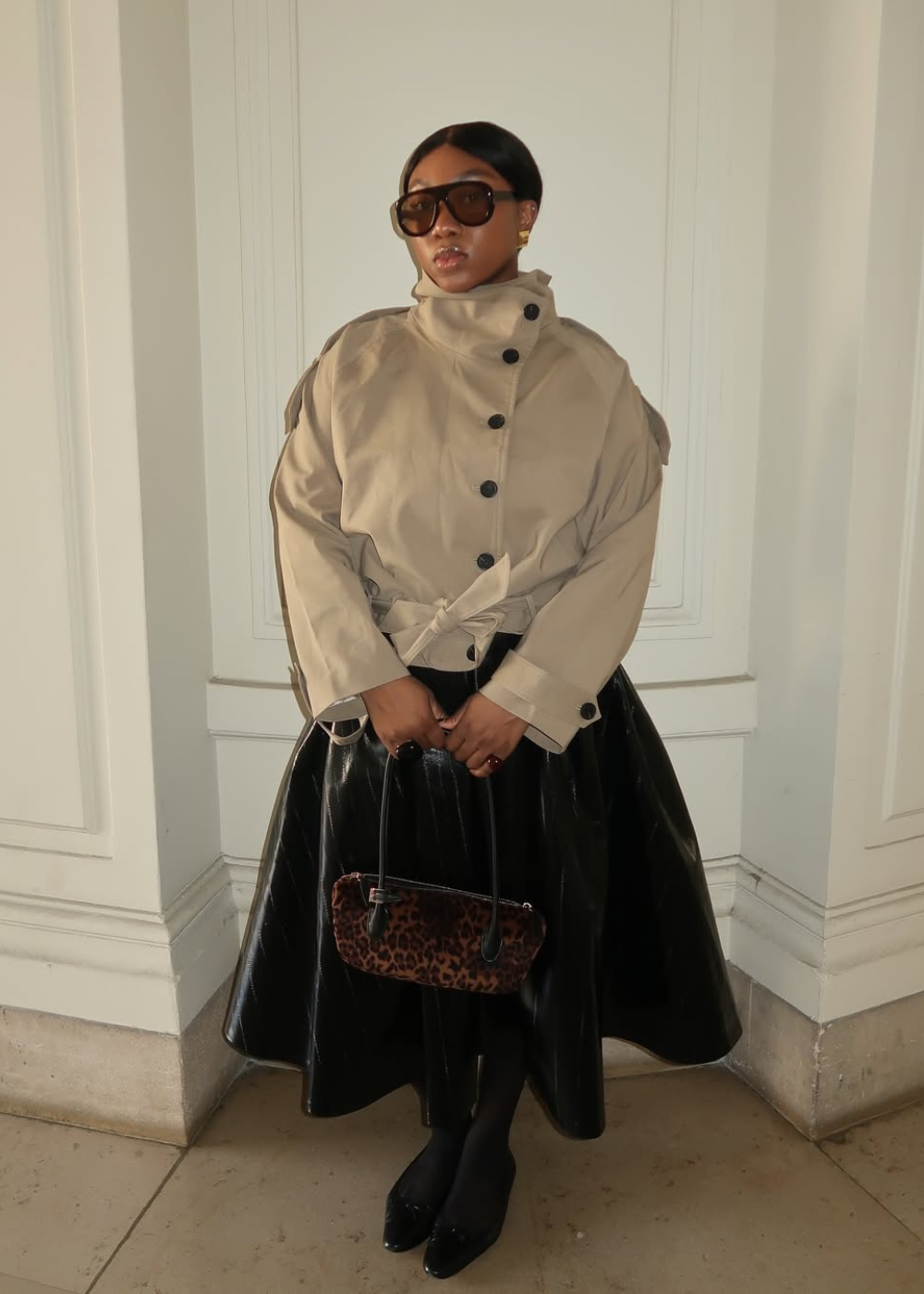 Modelo posa com óculos máscara oversized marrom, jaqueta bege estruturada e saia preta vinil, segurando bolsa animal print.