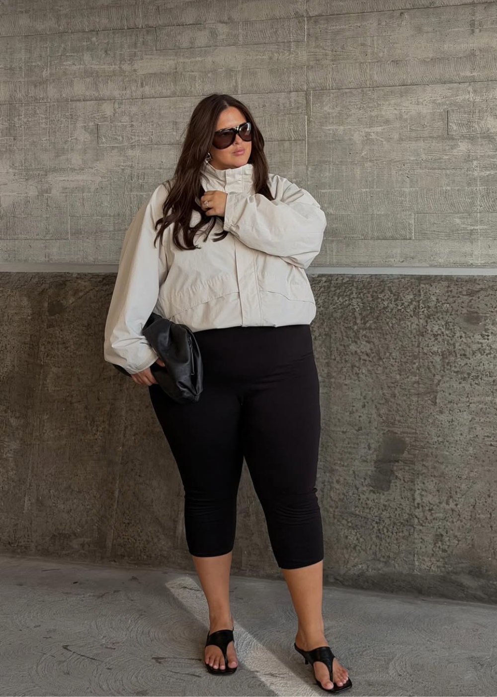 Modelo posa com óculos máscara escuros e oversized, jaqueta bege de tecido leve, legging preta justa e sandália preta.