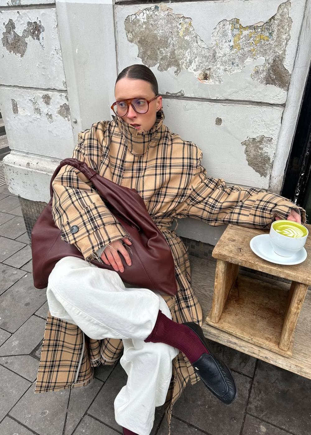 Modelo senta casualmente com óculos máscara de acetato marrom, sobretudo xadrez bege oversized, calça branca e bolsa vinho.
