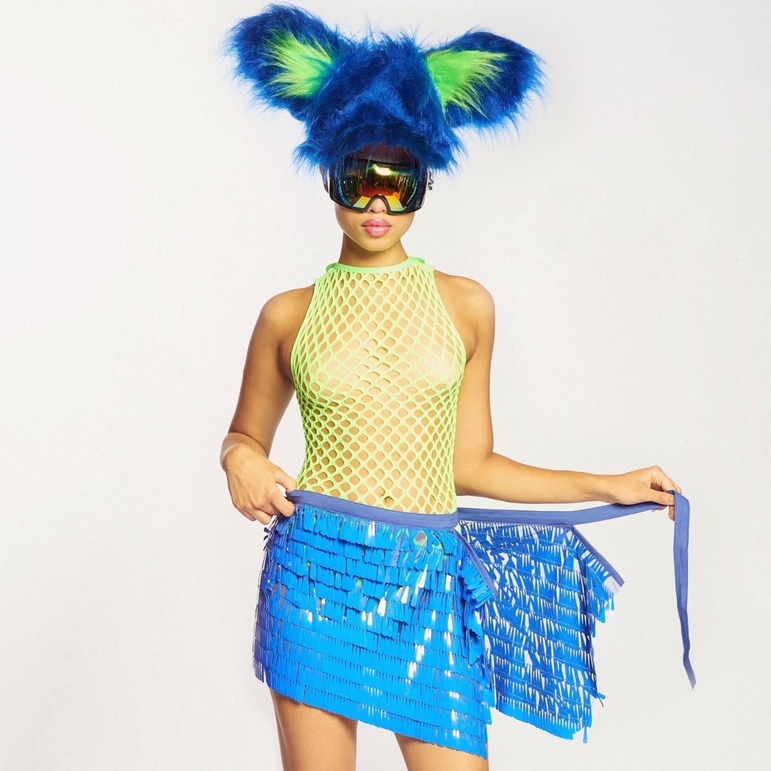 Modelo posa com body verde neon de tela, saia azul de franjas plásticas e chapéu felpudo azul com detalhes verdes, ideal