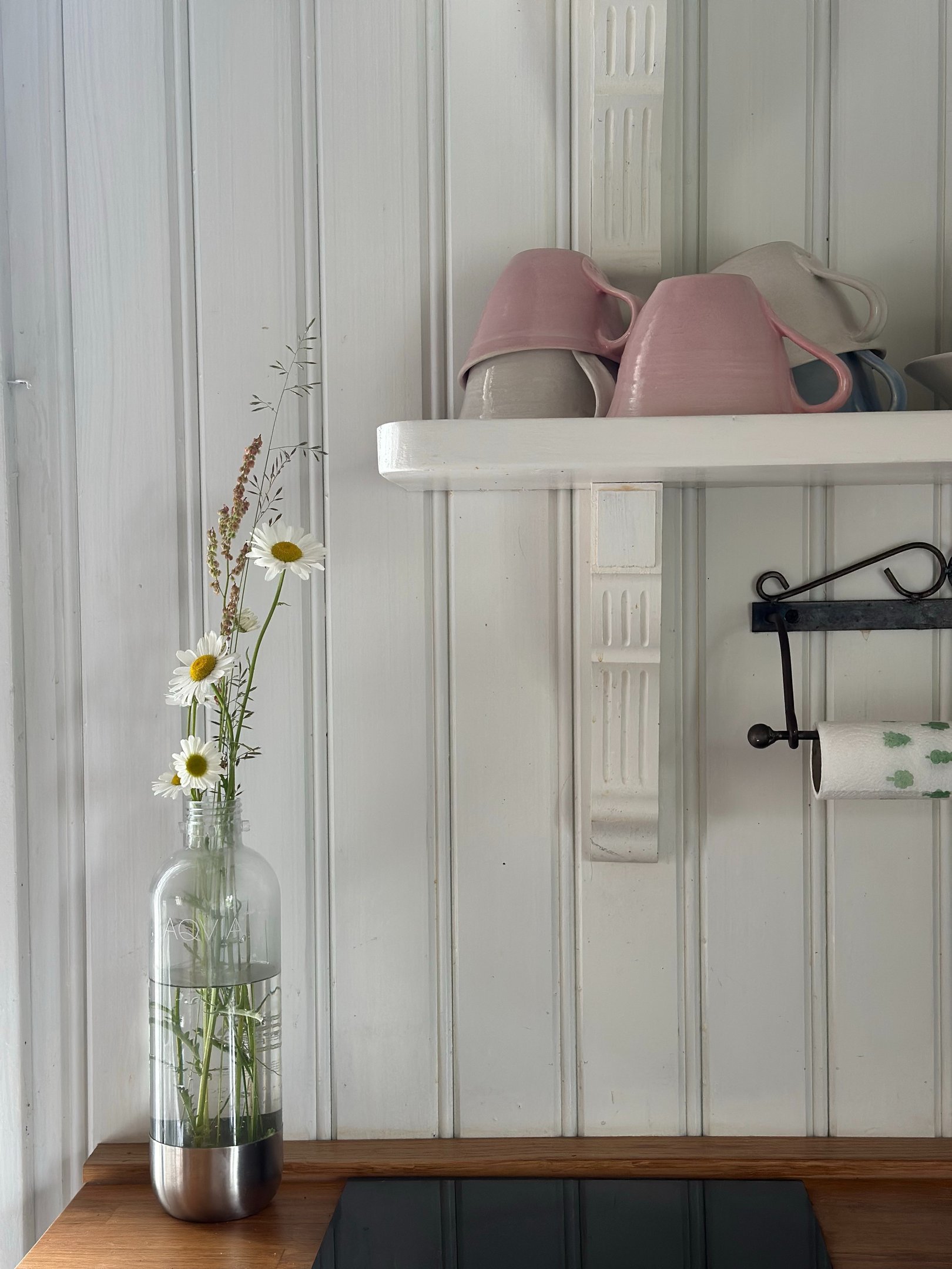 Cozinha retrô com prateleira branca de madeira, canecas em tons pastel e vaso de vidro com flores delicadas, clima acolhedor.