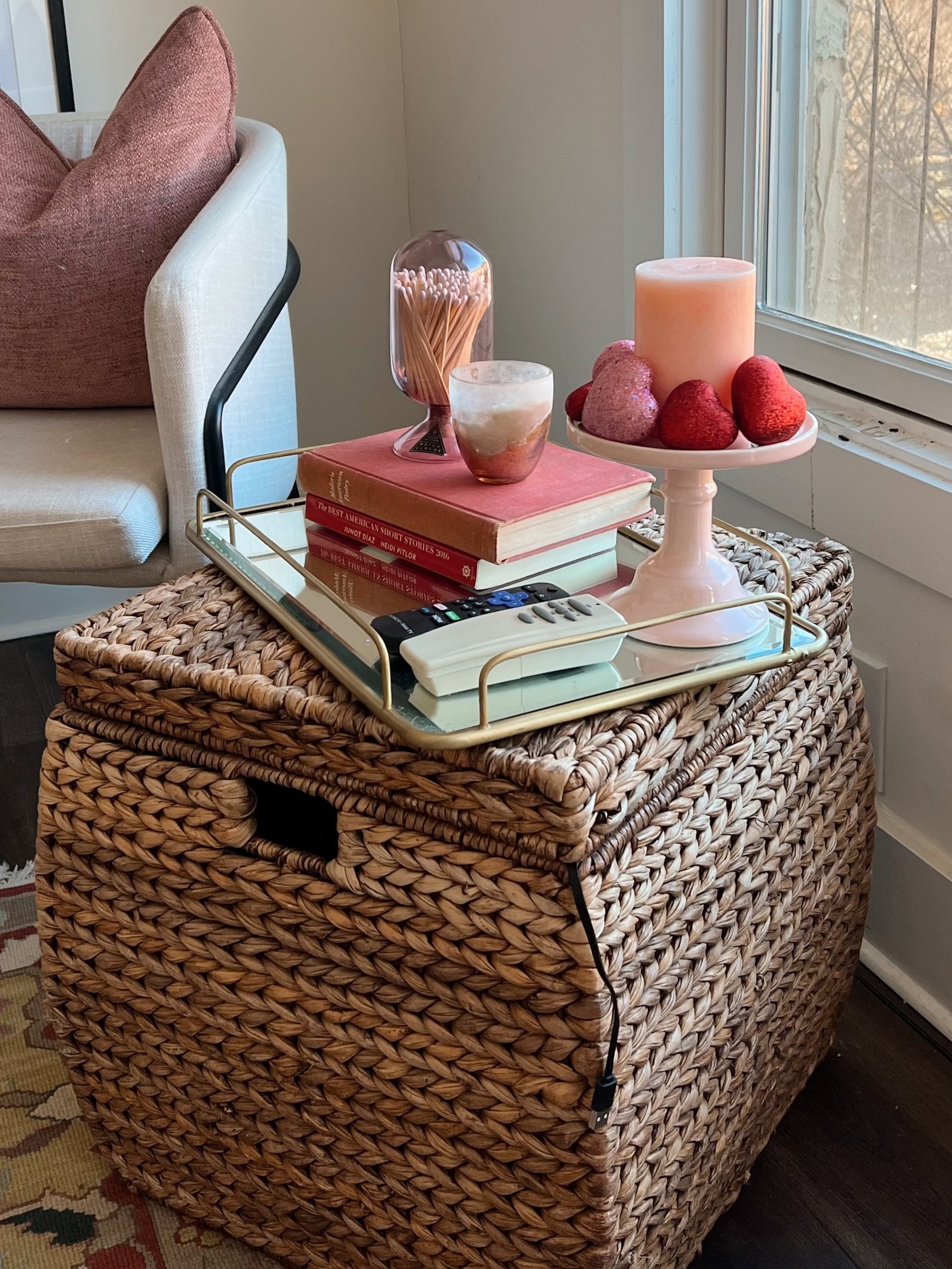 Cesto de fibra natural trançada funciona como mesa lateral com bandeja espelhada, livros, vela rosa e ambiente acolhedor