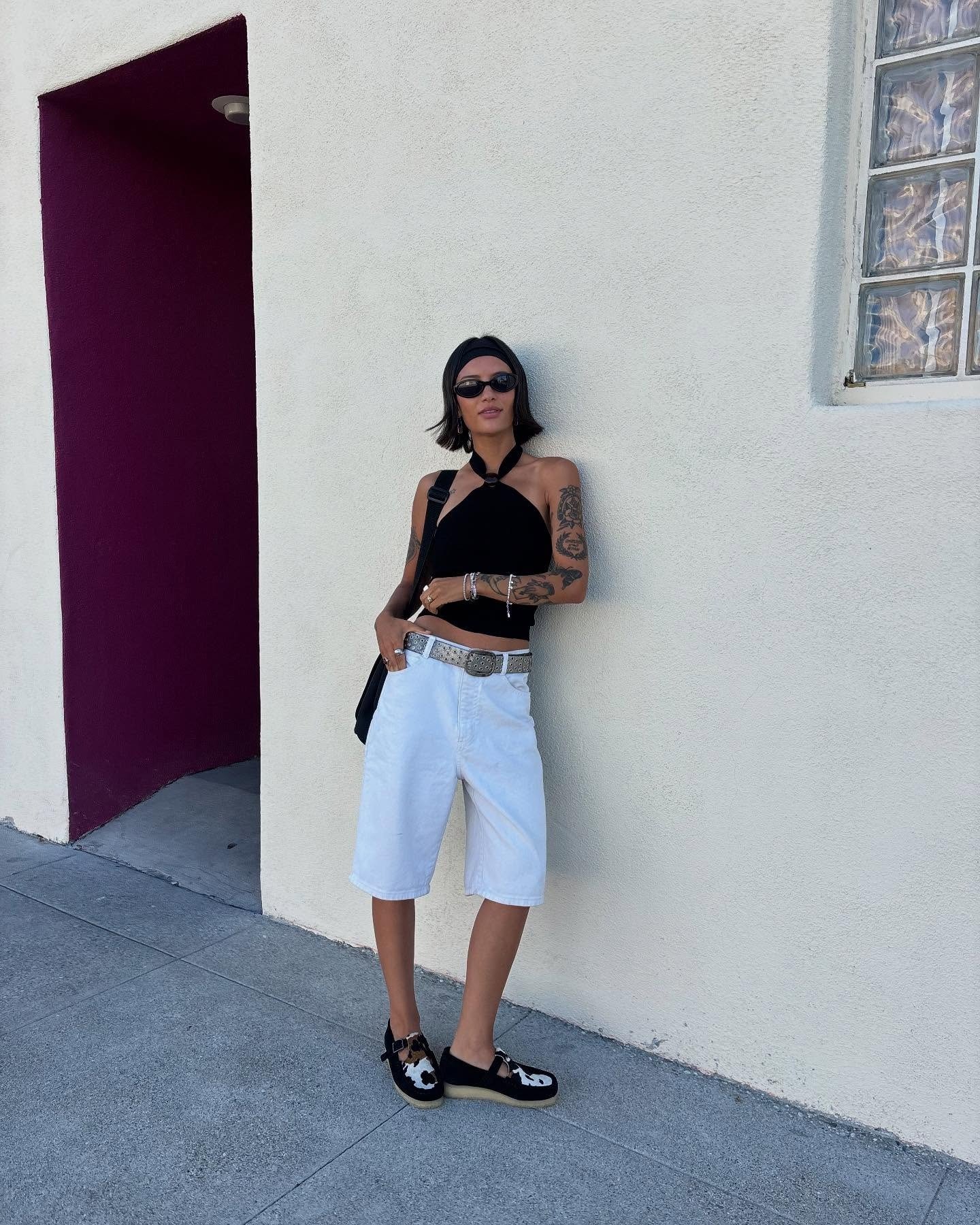 Modelo posa com top preto de amarração no pescoço em tecido leve, bermuda branca ampla, cinto prateado e sapatos com estampa