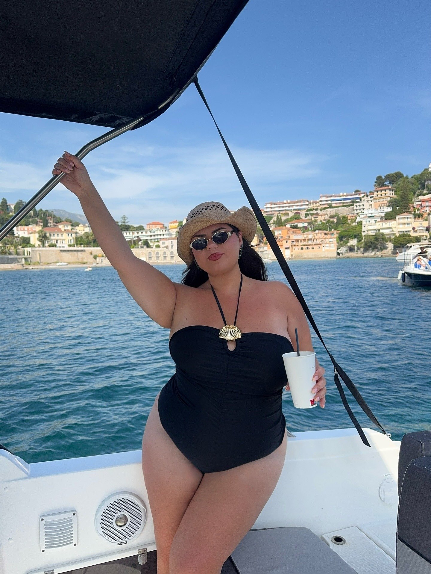 Modelo posa confiante em maiô preto com amarração no pescoço e pingente dourado, combinando chapéu de palha e óculos escuros