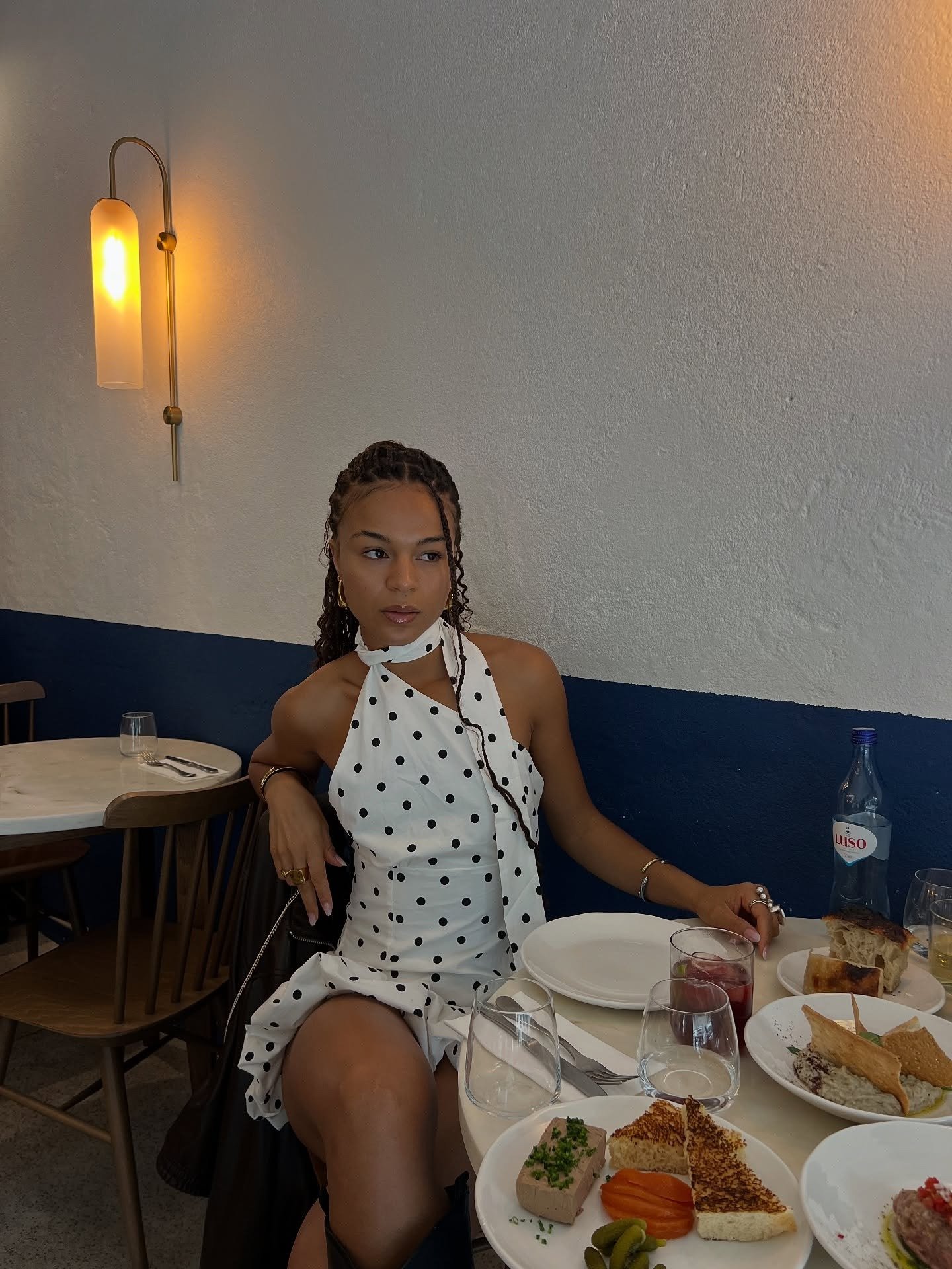 Modelo posa sentada com vestido branco de poás pretos, looks com amarração no pescoço e tecido leve