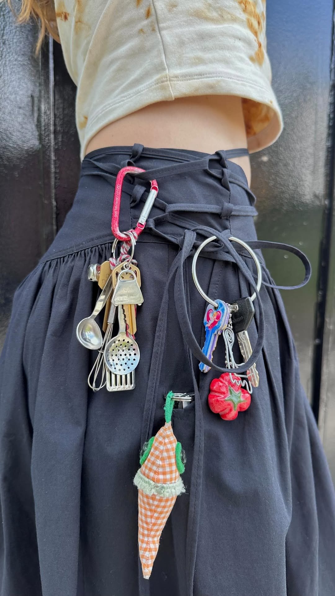 Saia preta com amarração frontal exibe pingentes para saias em metal e tecido, incluindo mini utensílios, chaveiro colorido