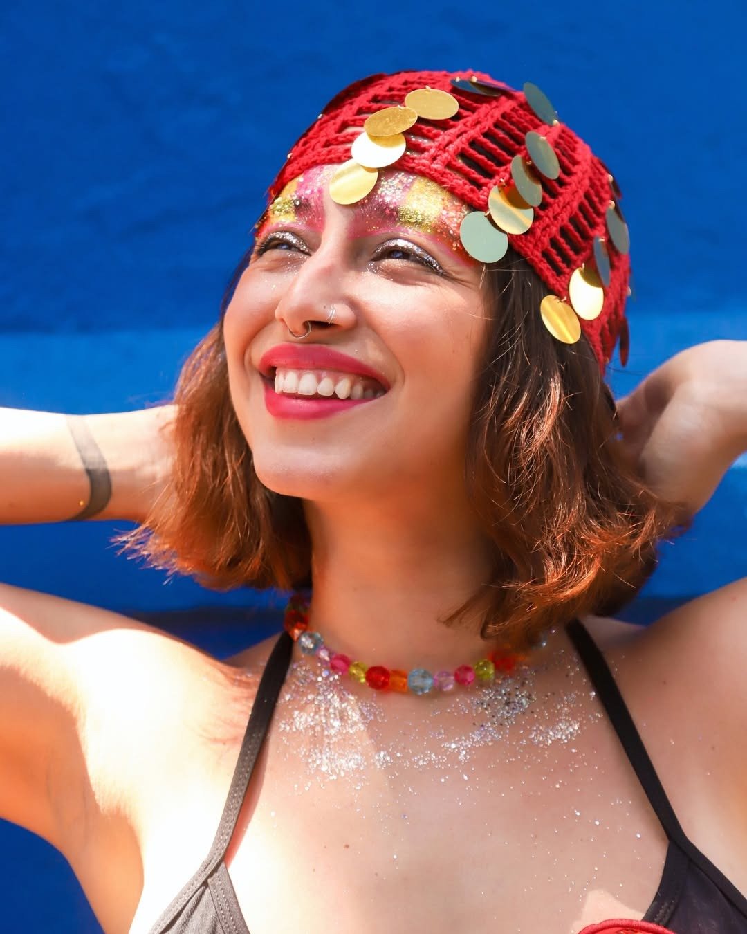 Mulher sorridente usa touca vermelha com paetês dourados, colar colorido de acrílico e brilho no corpo, ideal para lojas