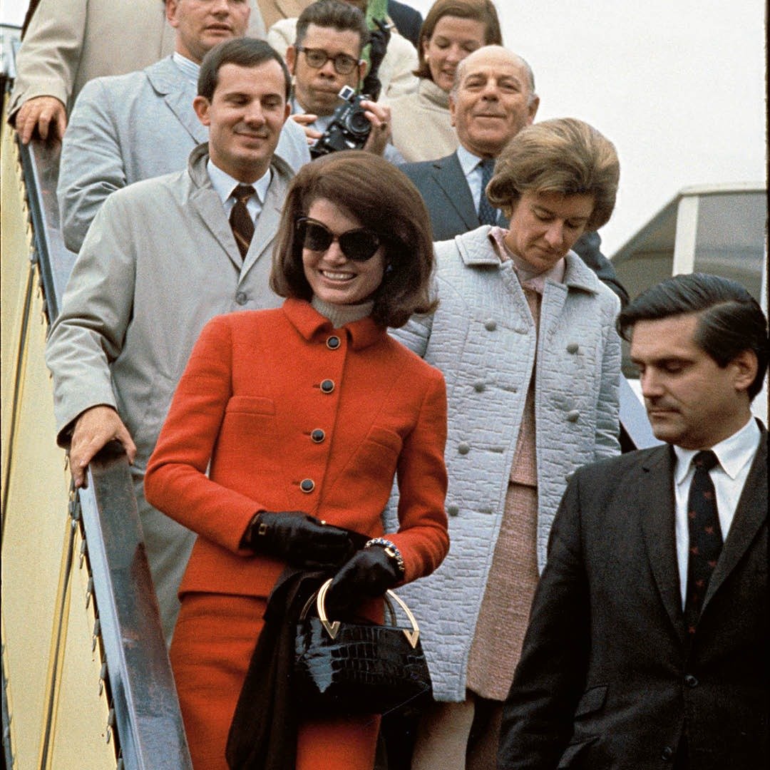 Modelo desce escada sorrindo com conjunto vermelho Valentino de alfaiataria, botões pretos, bolsa de couro croco e luvas