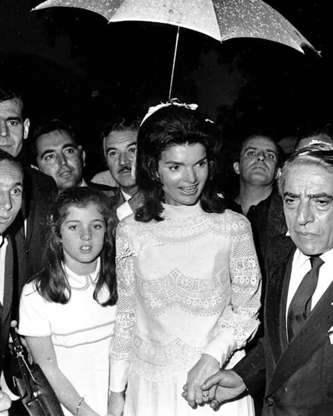 Valentino acompanha Jackie Kennedy em vestido branco de renda delicada, enquanto ela sorri segurando a mão de um homem sob