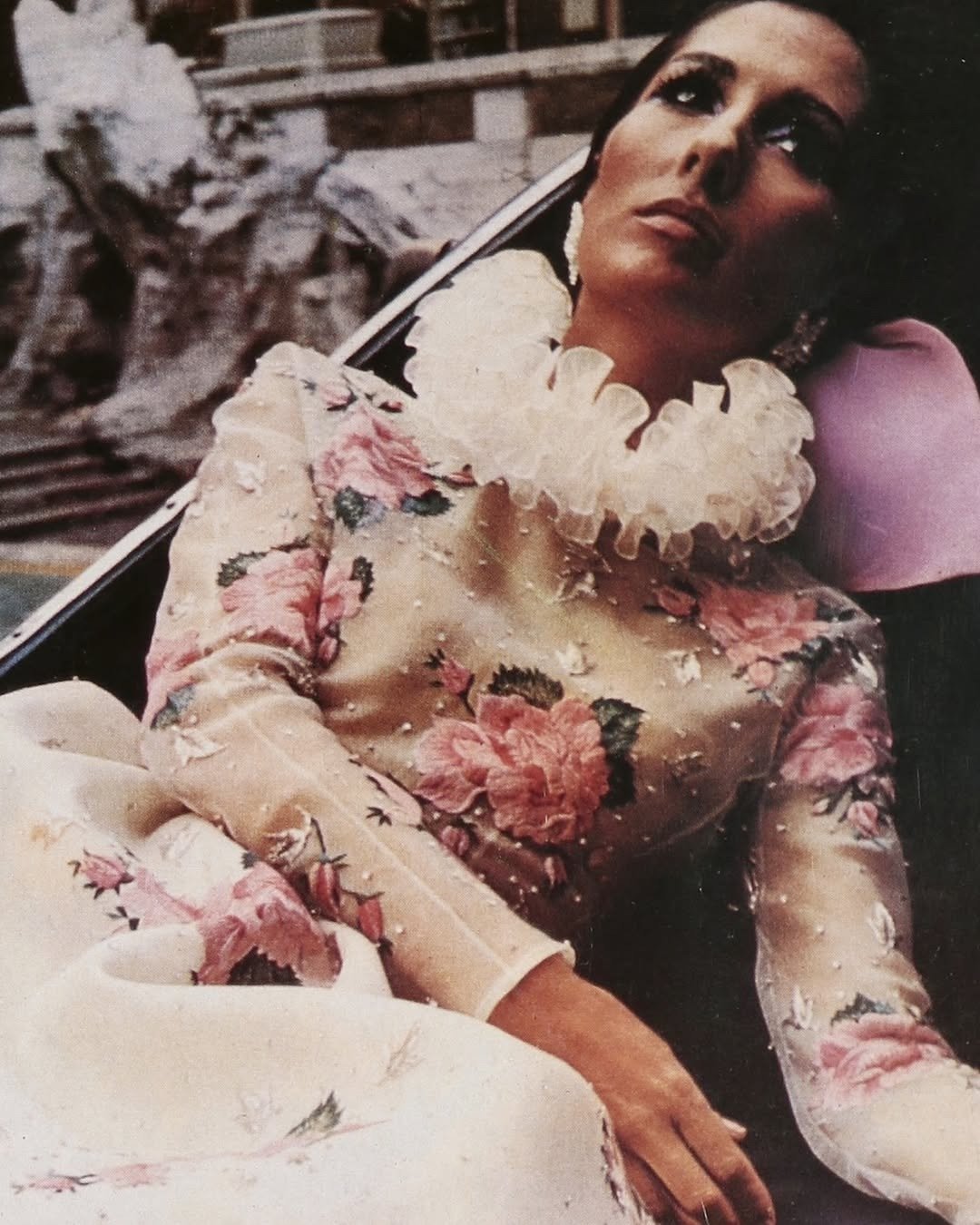 Modelo reclina com expressão sonhadora usando vestido Valentino de seda bordada com flores rosas e gola de tule volumosa, em