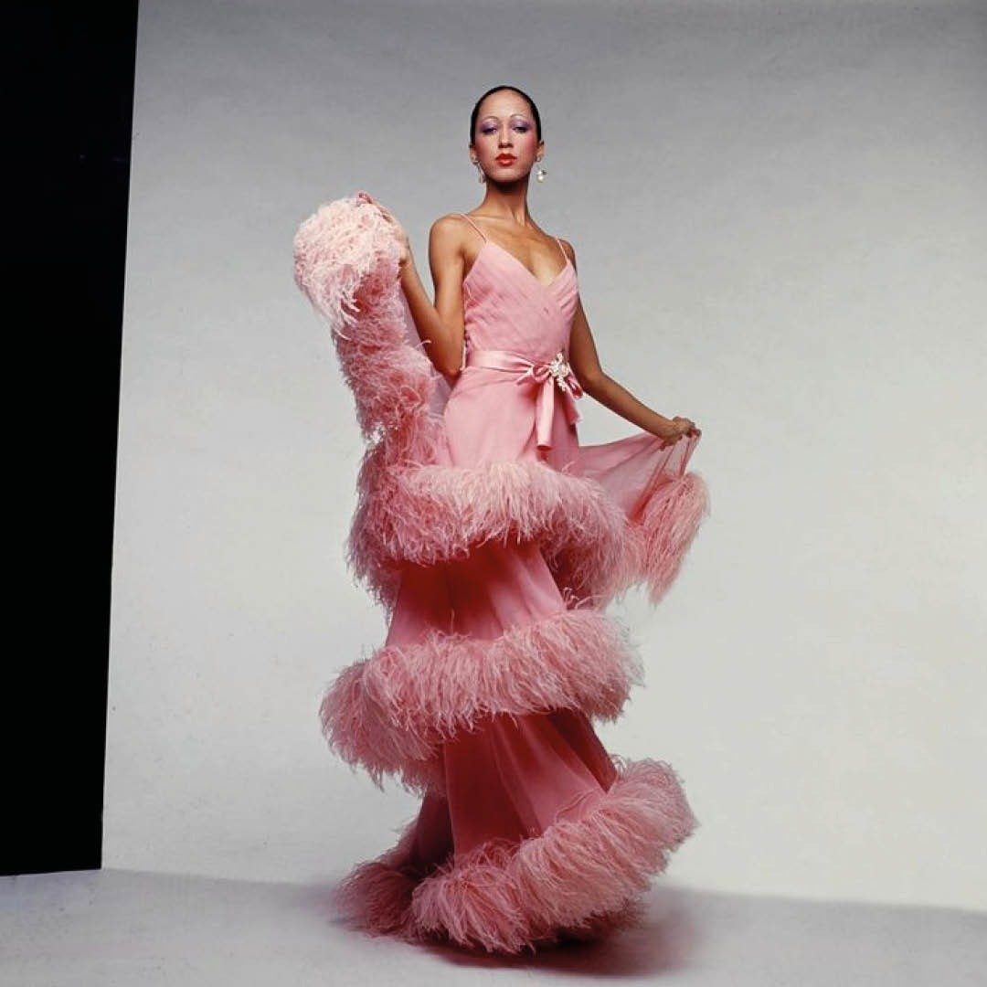 Modelo posa com vestido longo rosa Valentino em tecido fluido, detalhes em plumas e laço na cintura, exalando elegância e