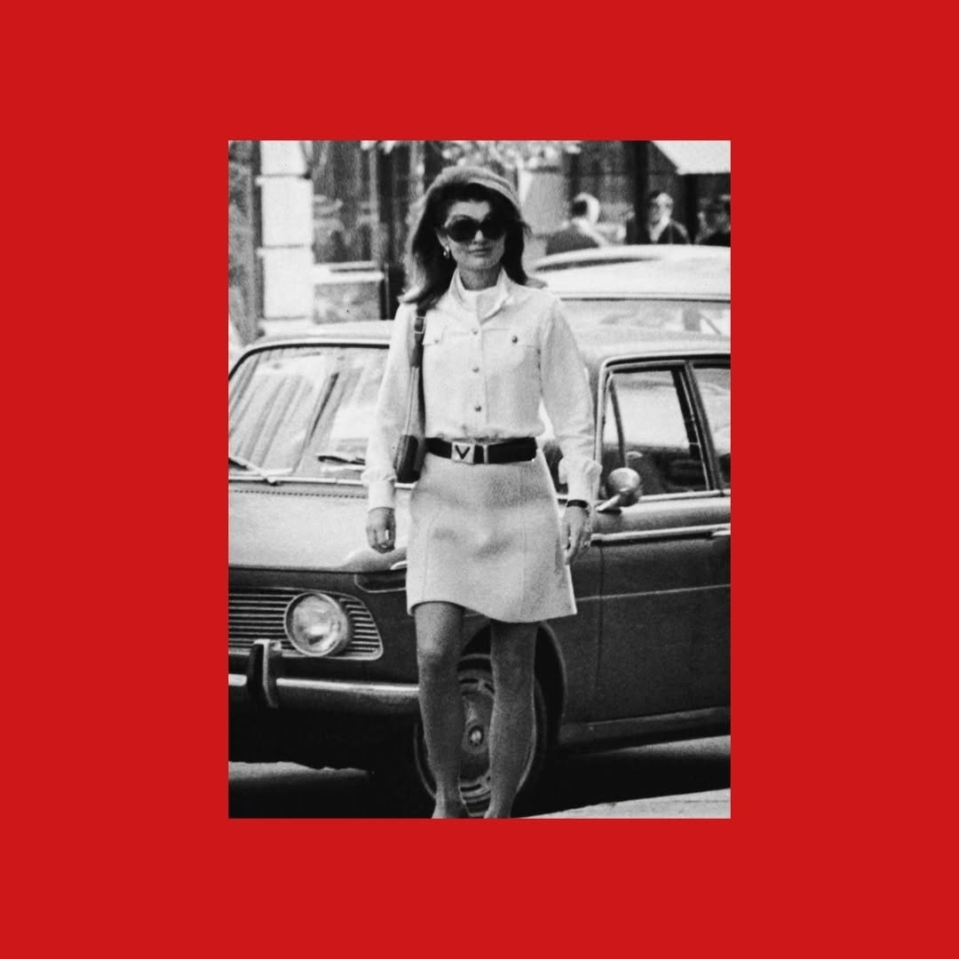 Modelo caminha confiante com camisa branca de botões, saia curta e cinto Valentino com logo em V, em cenário urbano vintage.