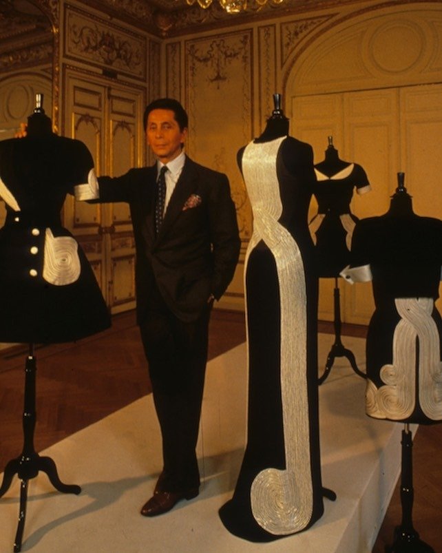 Valentino posa ao lado de vestidos pretos com bordados brancos em formas geométricas, destacando elegância clássica e