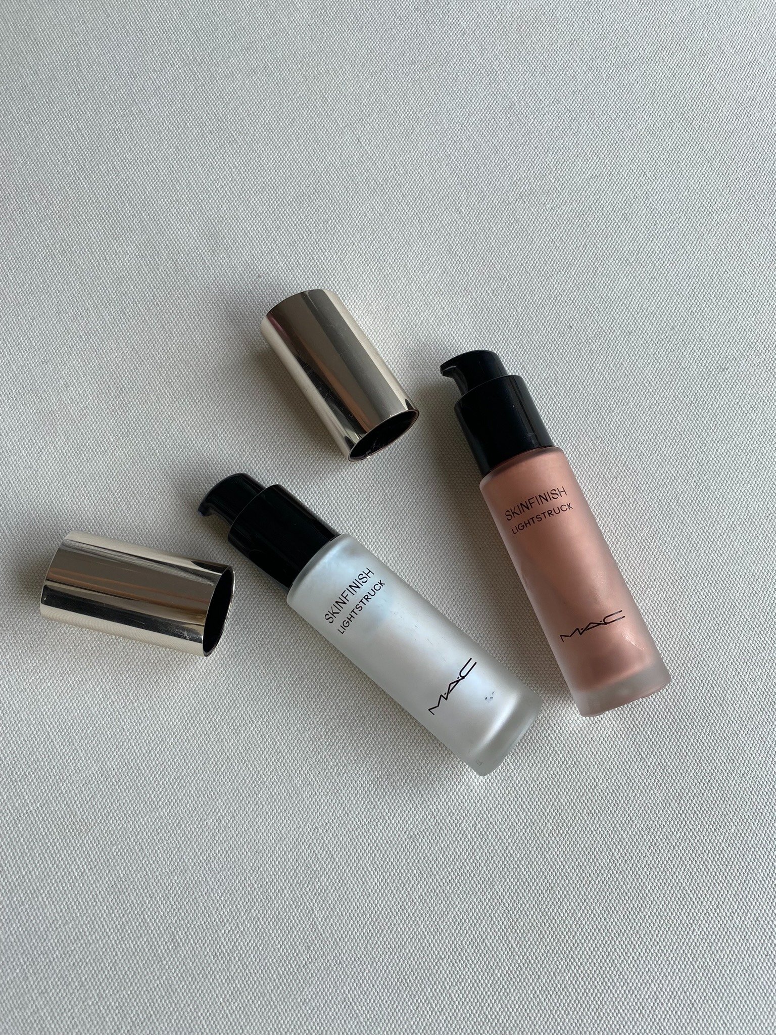 Frascos com pump de Skinfinish Lightstruck da MAC em tons perolado e bronze sobre tecido branco, destacando sofisticação
