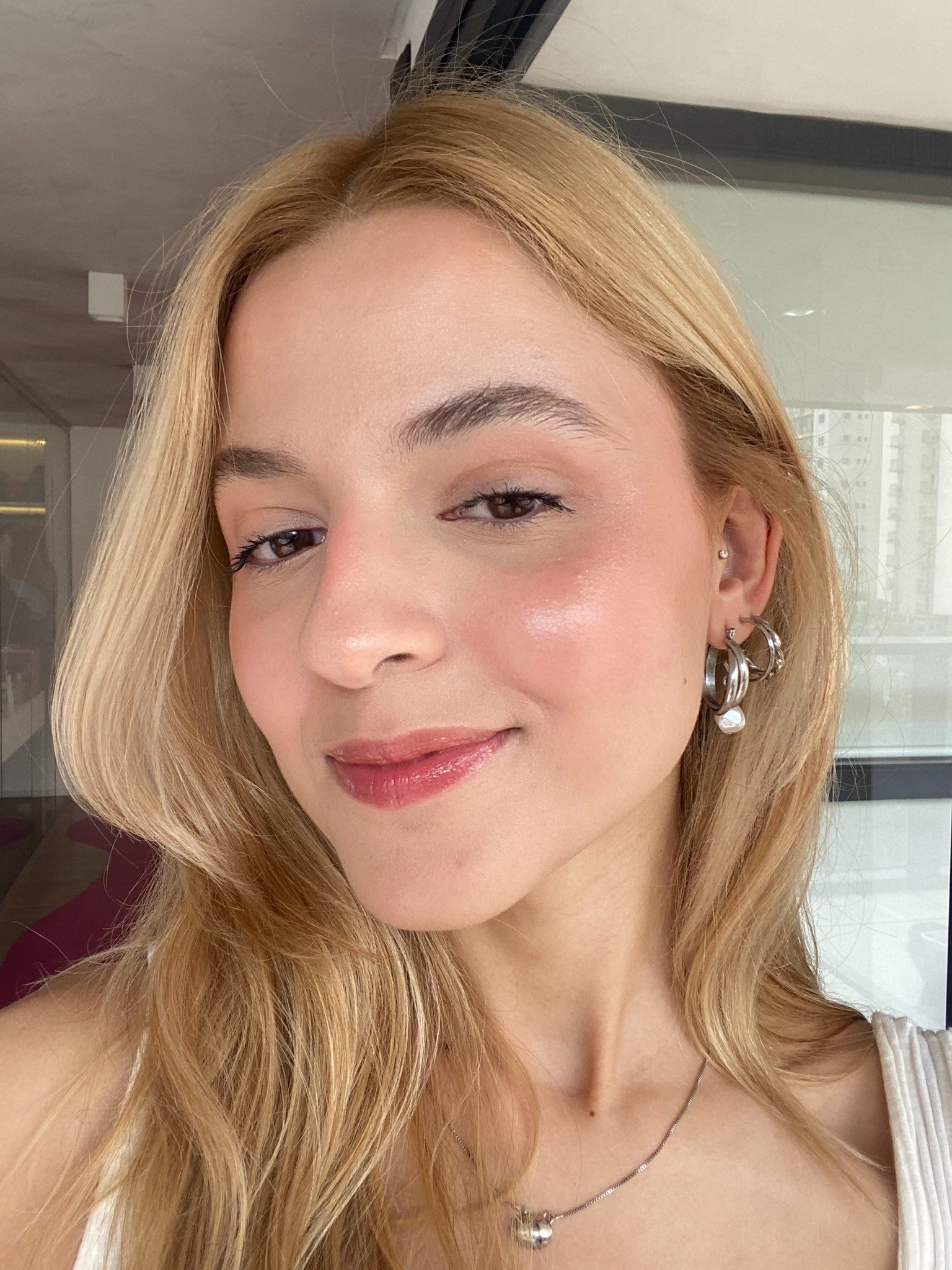 Modelo posa com pele iluminada pelo Skinfinish Lightstruck, cabelos loiros soltos, brincos prateados e blusa branca de alça