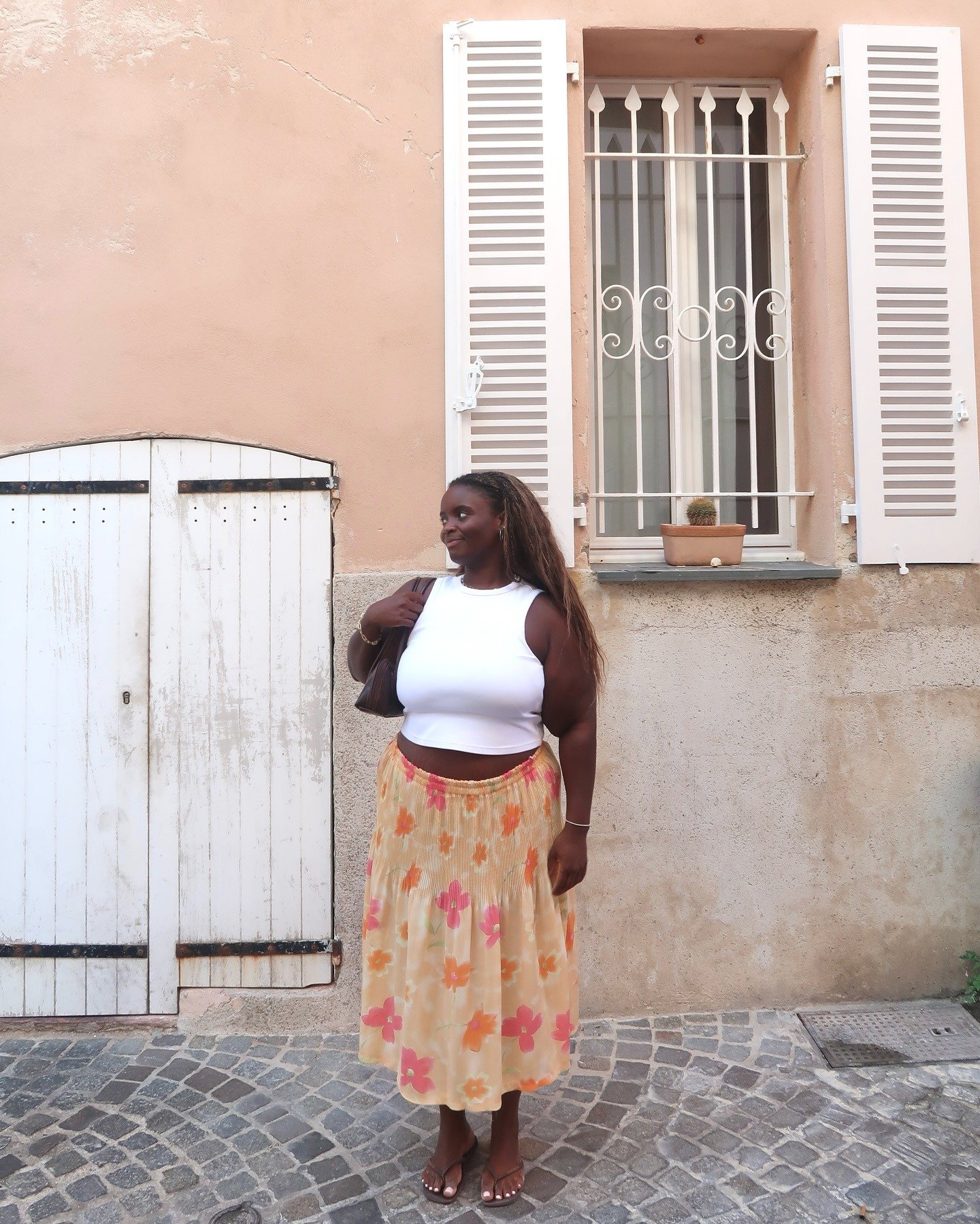 Modelo posa com roupa de calor composta por regata branca justa e saia midi plissada floral em tons pastel, em clima leve e