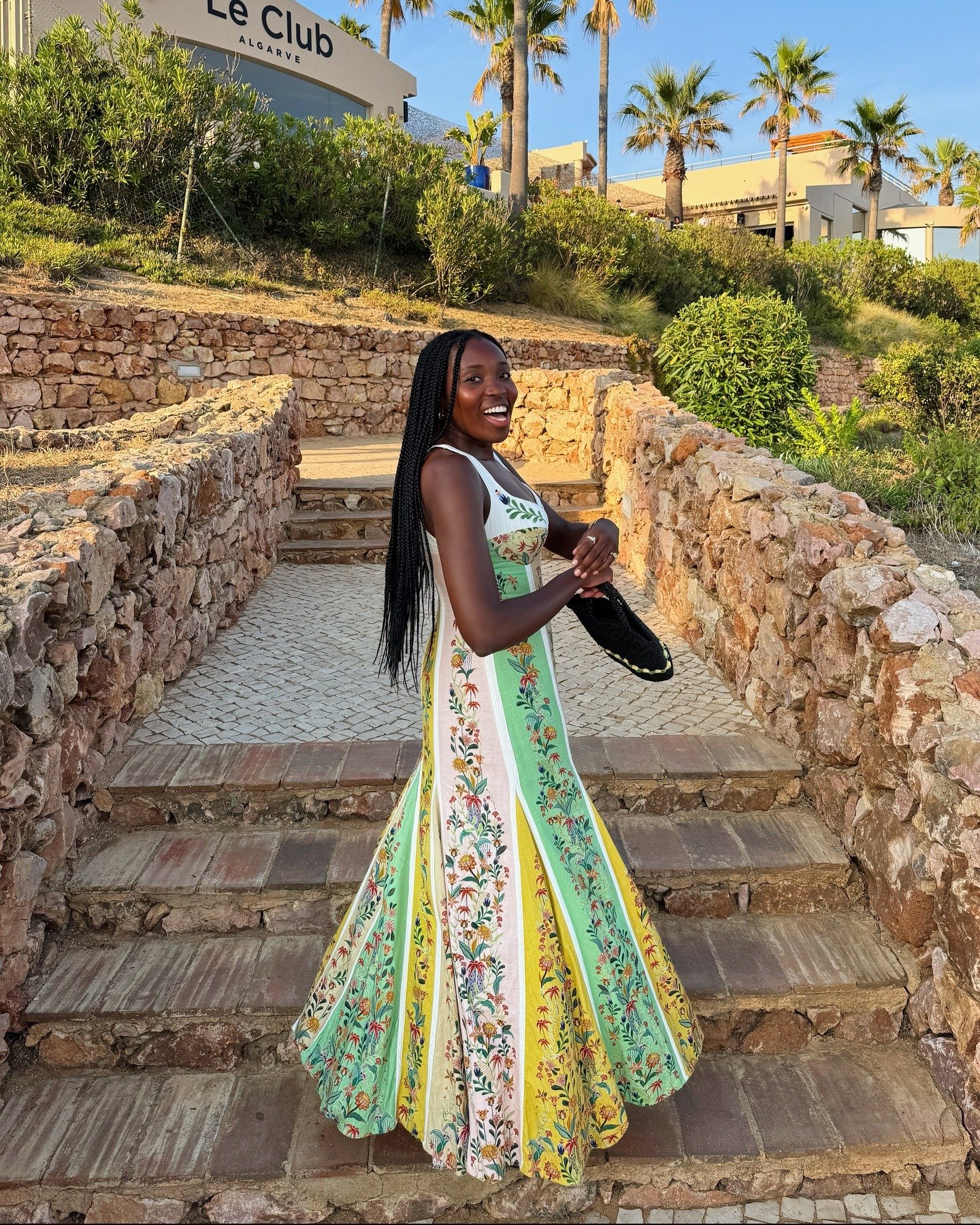 Modelo sorri e posa com vestido longo de tecido leve em tons verde, amarelo e branco com estampa floral, ideal para roupa de