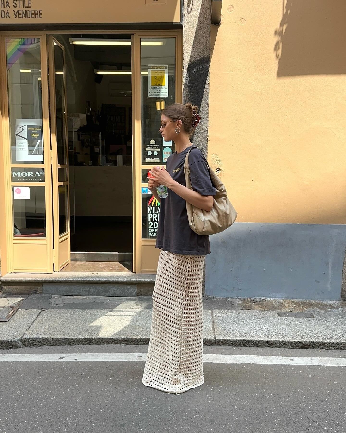 Modelo posa de perfil na rua com saia de crochê bege longa, camiseta preta oversized e bolsa bege casual, em clima urbano