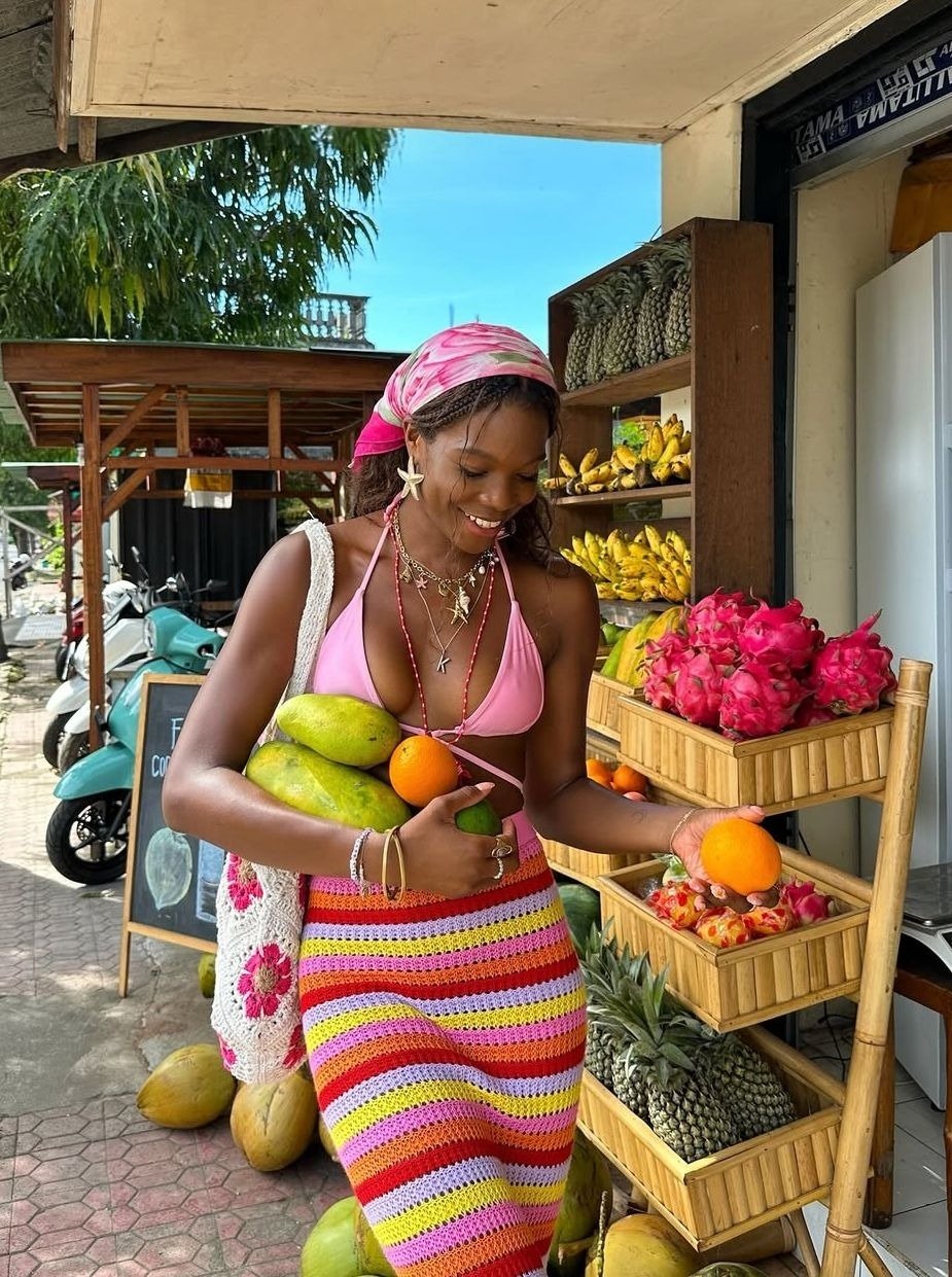 Modelo sorri segurando frutas, veste saia de crochê listrada em rosa, amarelo e vermelho com top rosa e lenço na cabeça.