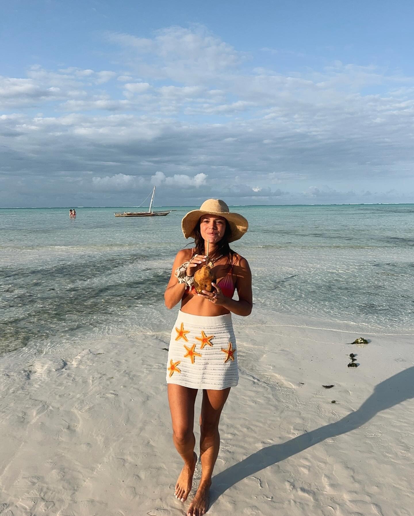 Modelo caminha na praia com saia de crochê branca decorada por estrelas do mar laranja, chapéu de palha e clima descontraído.
