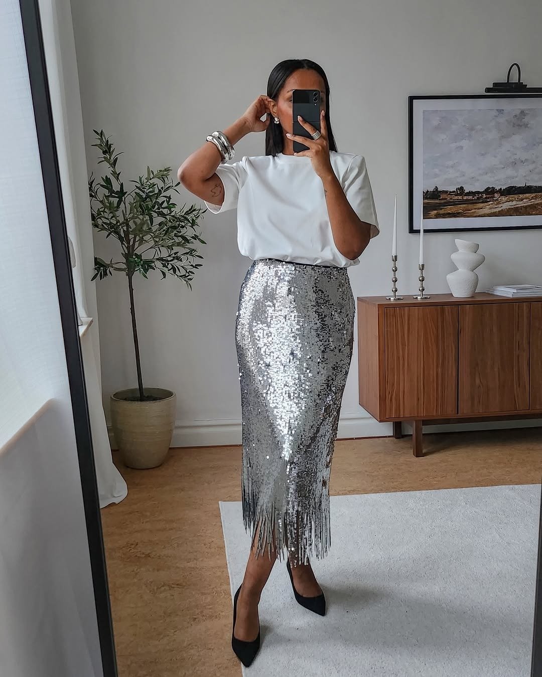 Modelo posa em saia de paetê prateada com franjas, combinada com blusa branca de manga curta e scarpin preto.