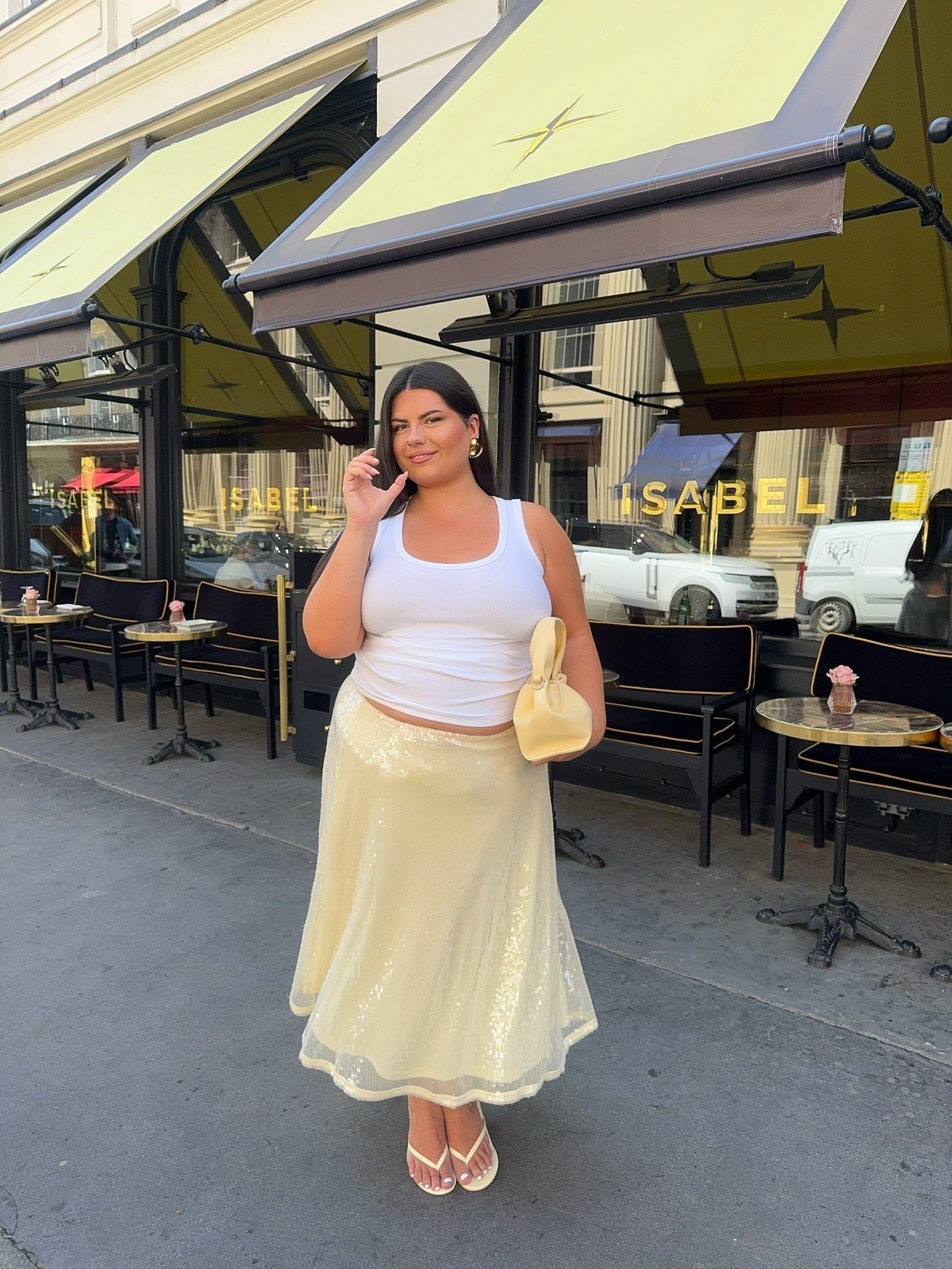 Modelo posa confiante com saia de paetê amarela midi fluida, regata branca e bolsa bege em cenário urbano ensolarado.