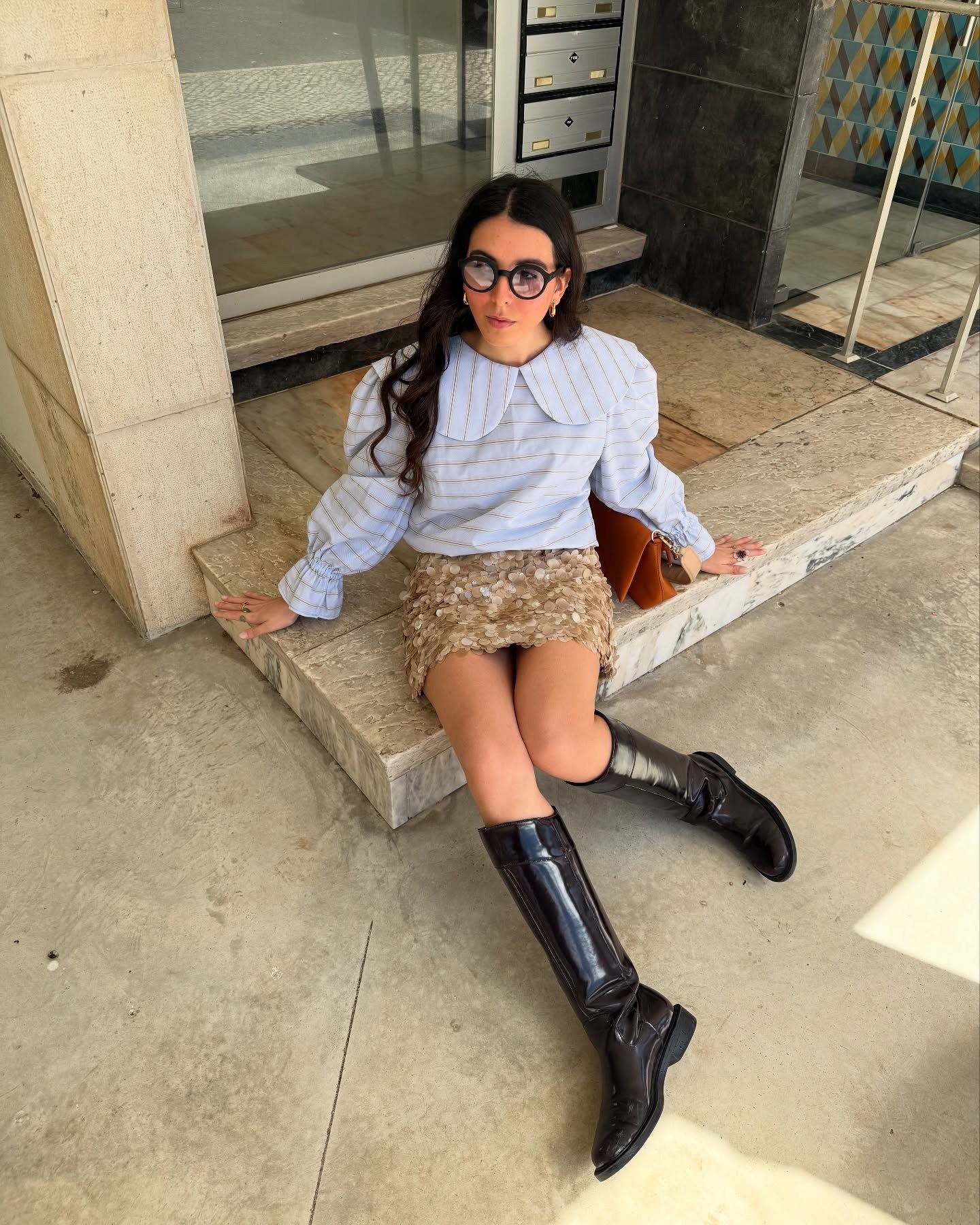 Modelo senta casualmente com saia de paetê dourada curta, blusa branca com gola oversized e botas pretas de cano alto.