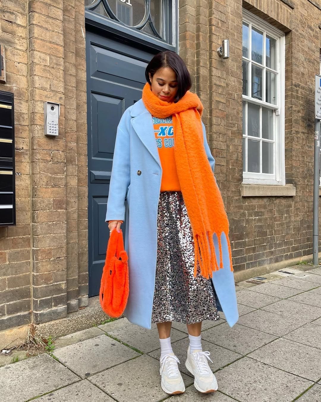 Modelo posa olhando para baixo com saia de paetê prata midi, casaco azul claro, moletom e cachecol laranja vibrante