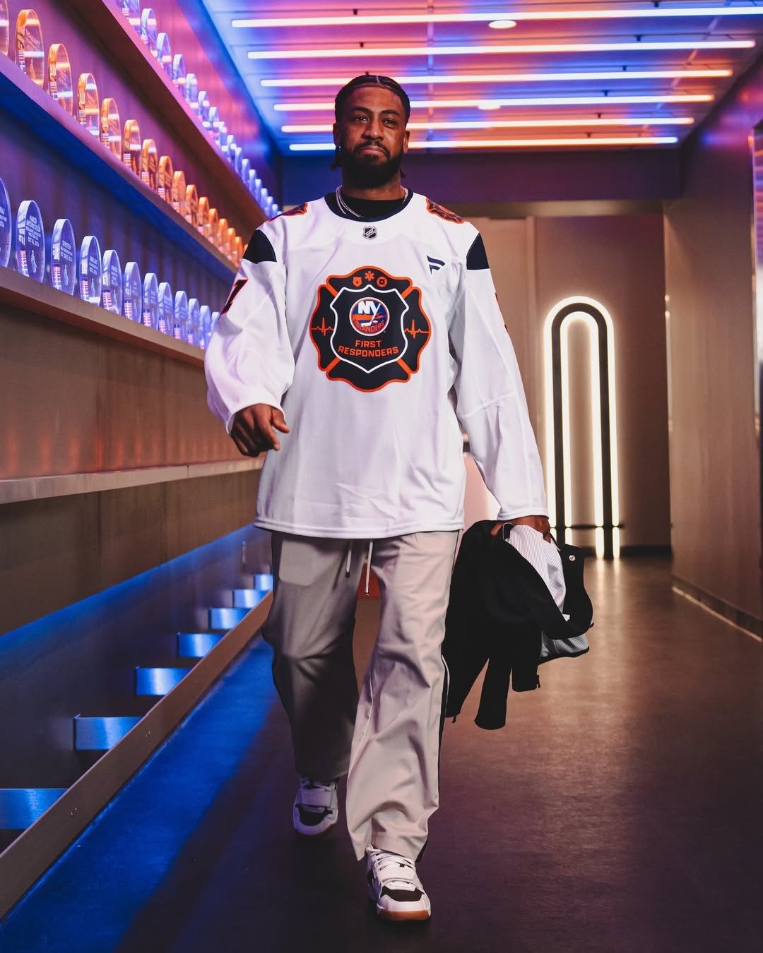 Homem caminha confiante em corredor iluminado, vestindo camiseta de hóquei branca com logo laranja e preto.