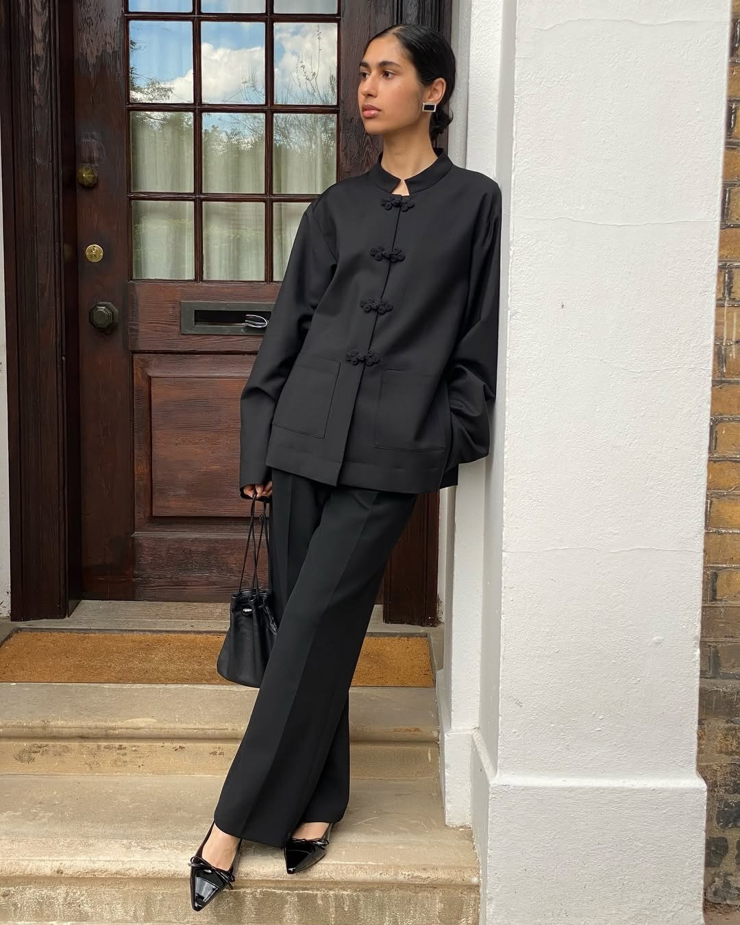 Modelo posa encostada em parede com conjunto preto de alfaiataria fluida, jaqueta com nó chinês e sapatos de bico fino