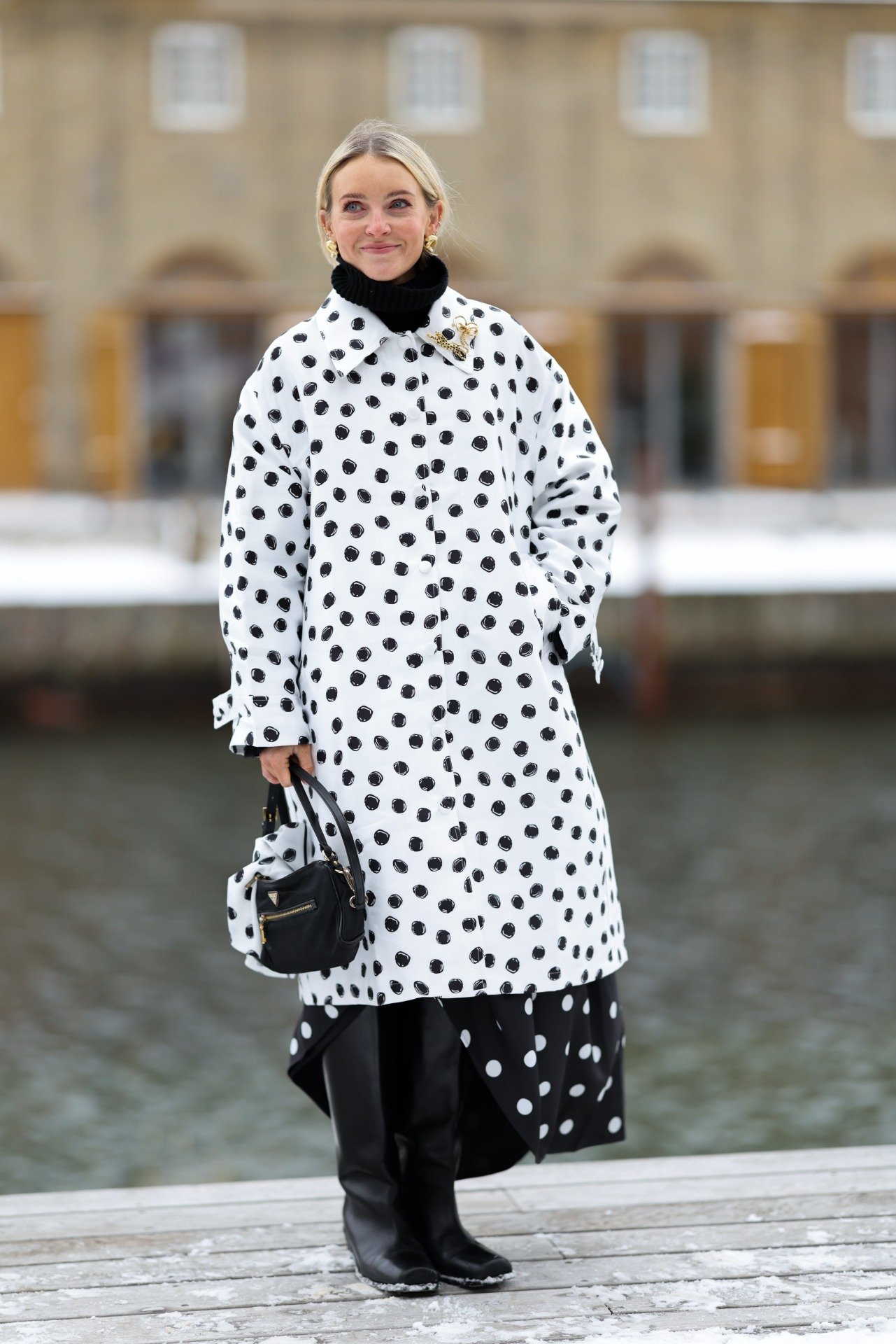 Na Semana de Moda de Copenhagen, modelo posa com casaco branco de poás pretos, saia assimétrica, botas pretas e bolsa