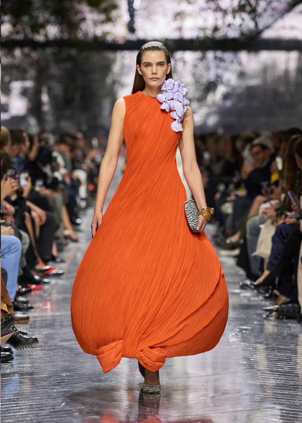 Modelo caminha confiante com vestido laranja plissado de modelagem volumosa e detalhe floral lilás no ombro, evidenciando