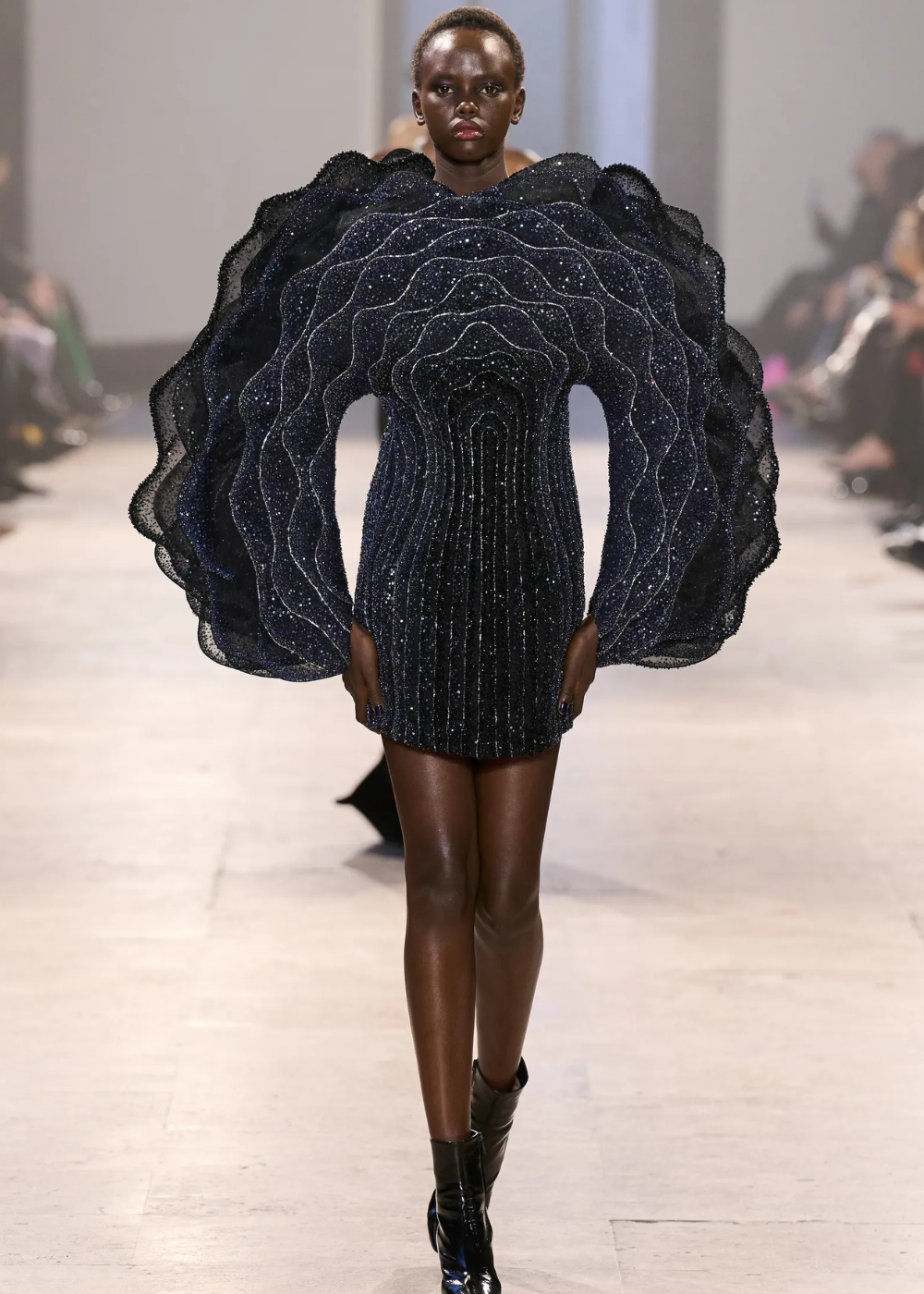 Modelo desfila com vestido curto estruturado em camadas onduladas de tule preto brilhante, destacando as tendências da