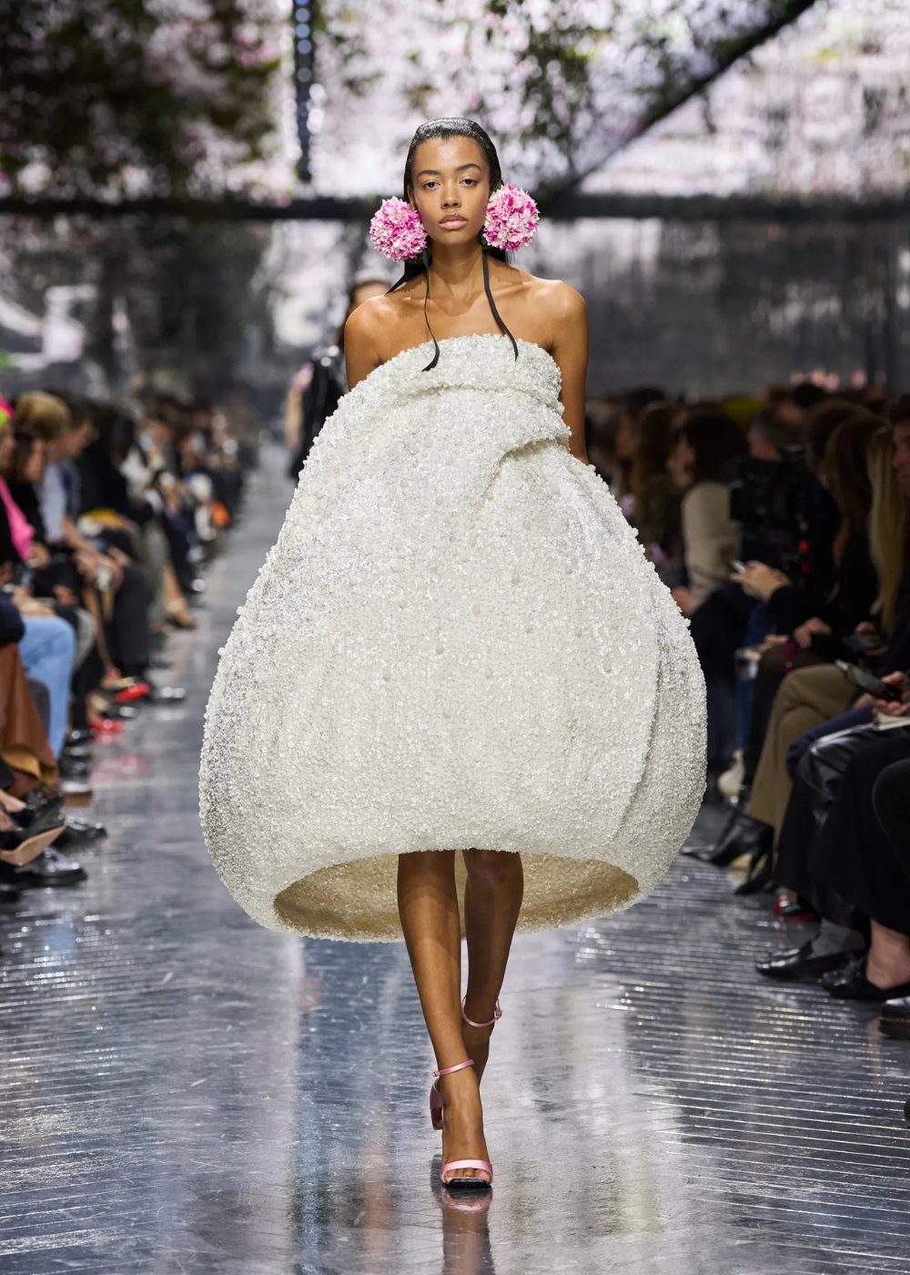 Modelo caminha confiante com vestido volumoso branco texturizado em formato arredondado, combinando maxi acessórios florais