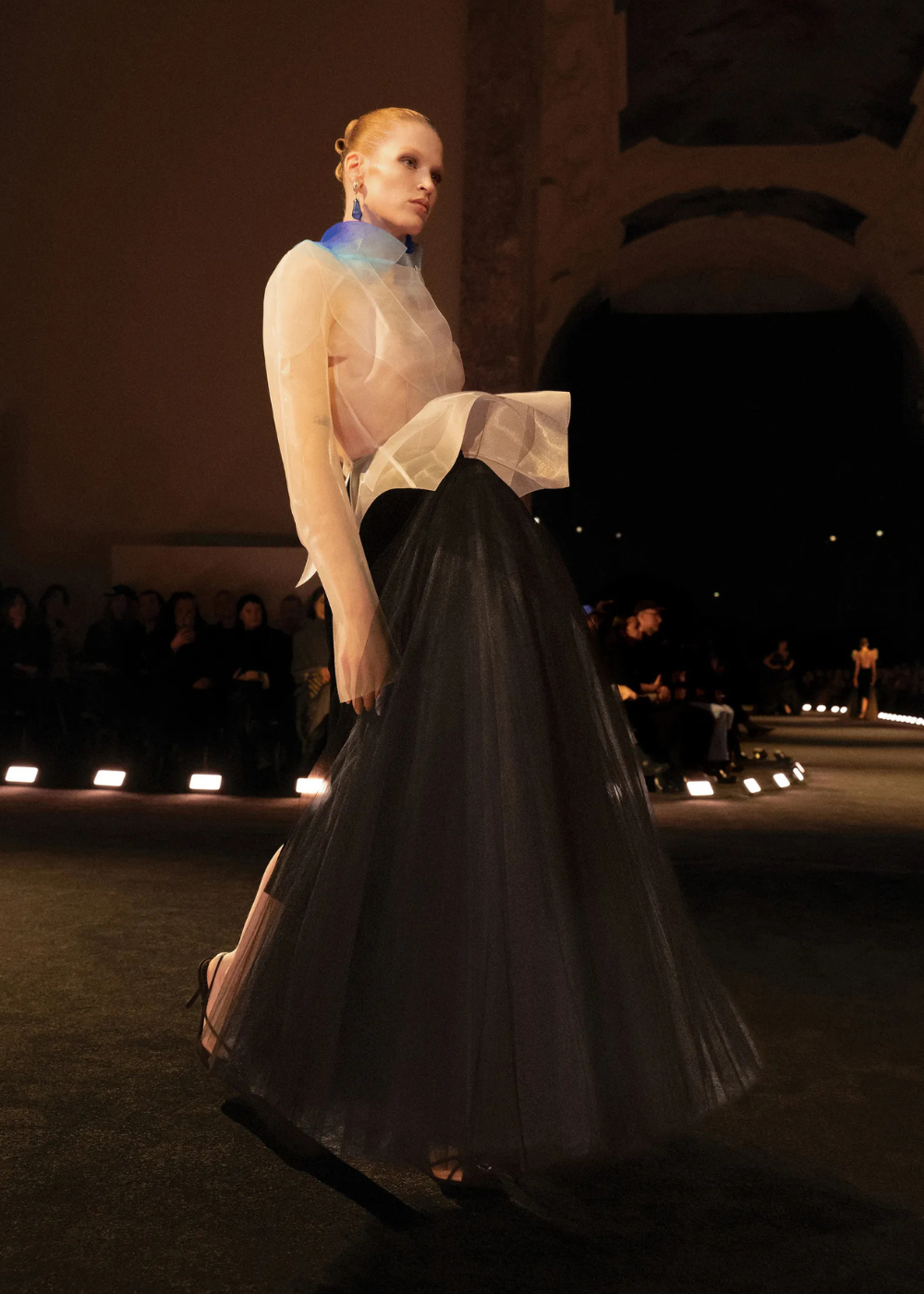 Modelo posa com blusa transparente de organza com gola azul e saia preta volumosa em tule, refletindo as tendências da