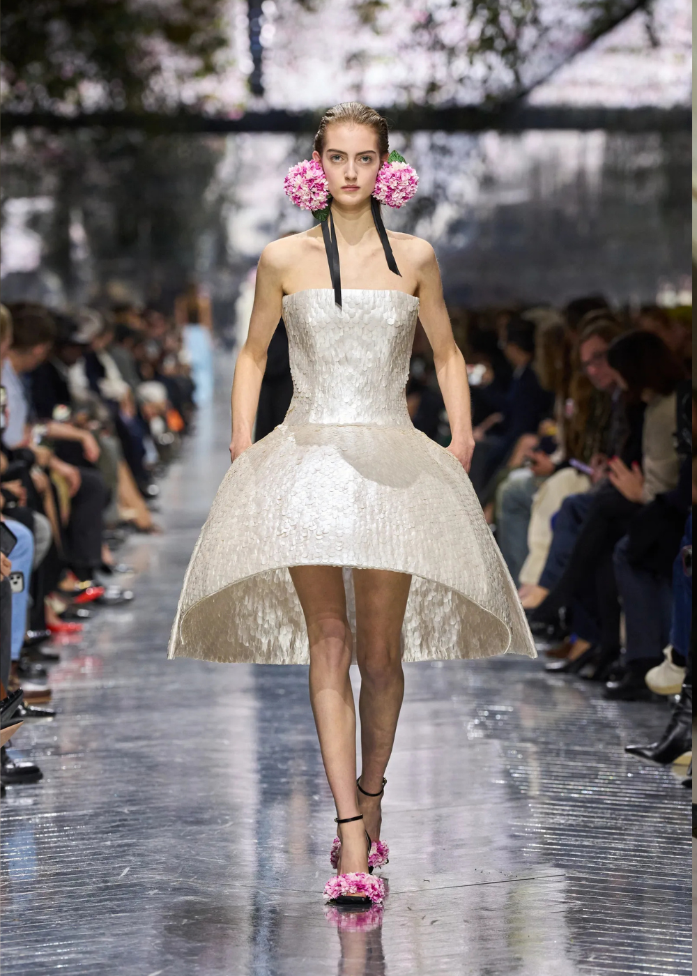 Modelo caminha com vestido tomara que caia estruturado em tecido texturizado off-white e acessórios florais vibrantes,