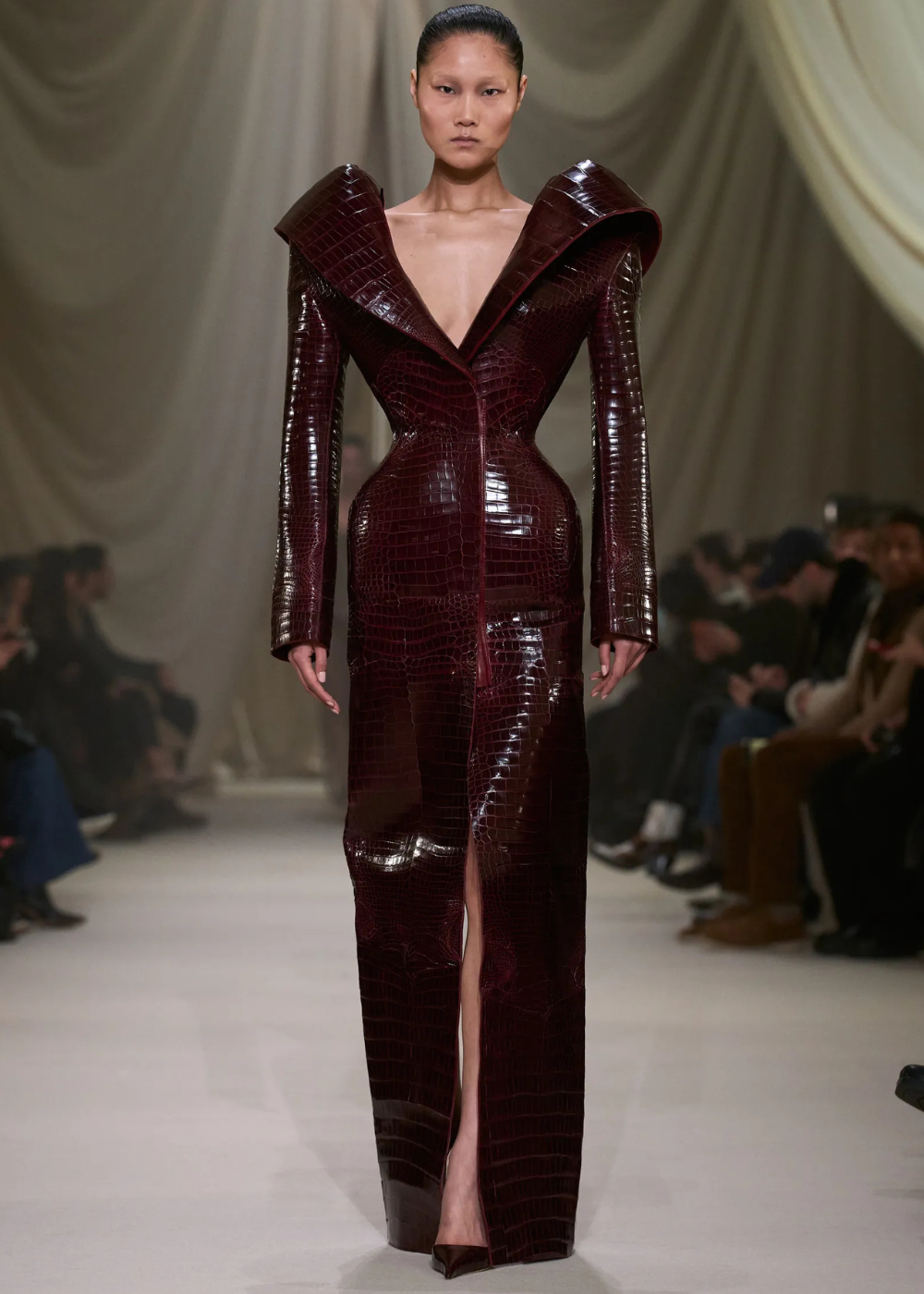 Modelo posa com vestido longo estruturado em couro verniz texturizado vinho, decote profundo e fenda frontal, refletindo