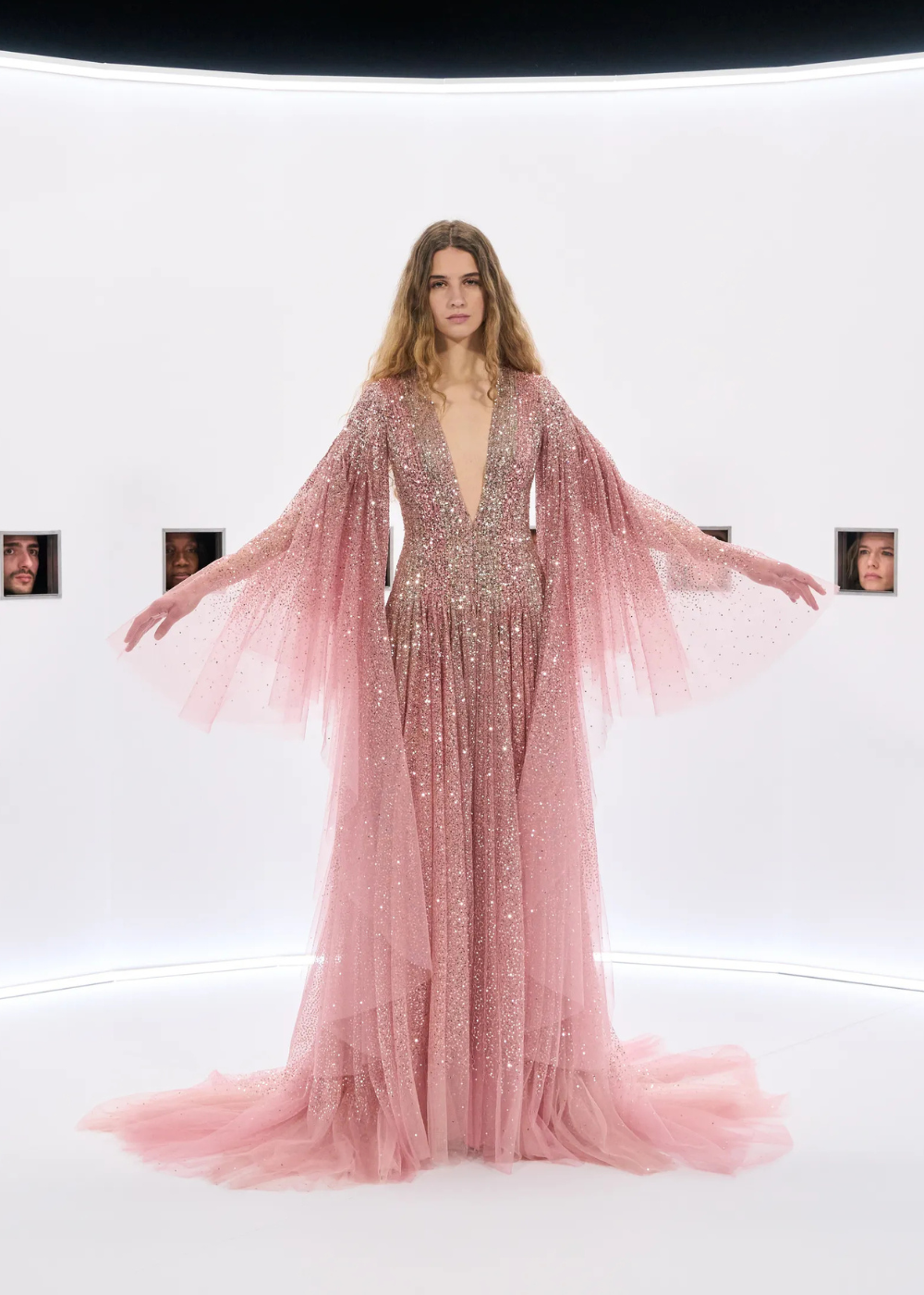 Modelo posa com vestido longo rosa em tule brilhante, mangas amplas e decote profundo, refletindo a delicadeza e