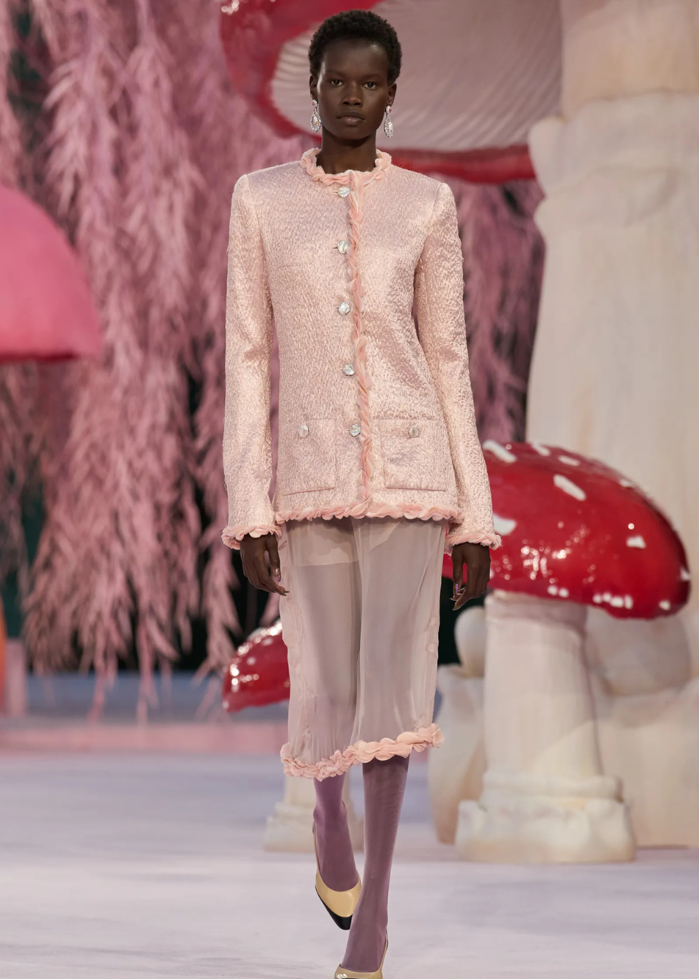 Modelo caminha com postura confiante usando blazer rosa texturizado e saia transparente com detalhes em babados, destacando