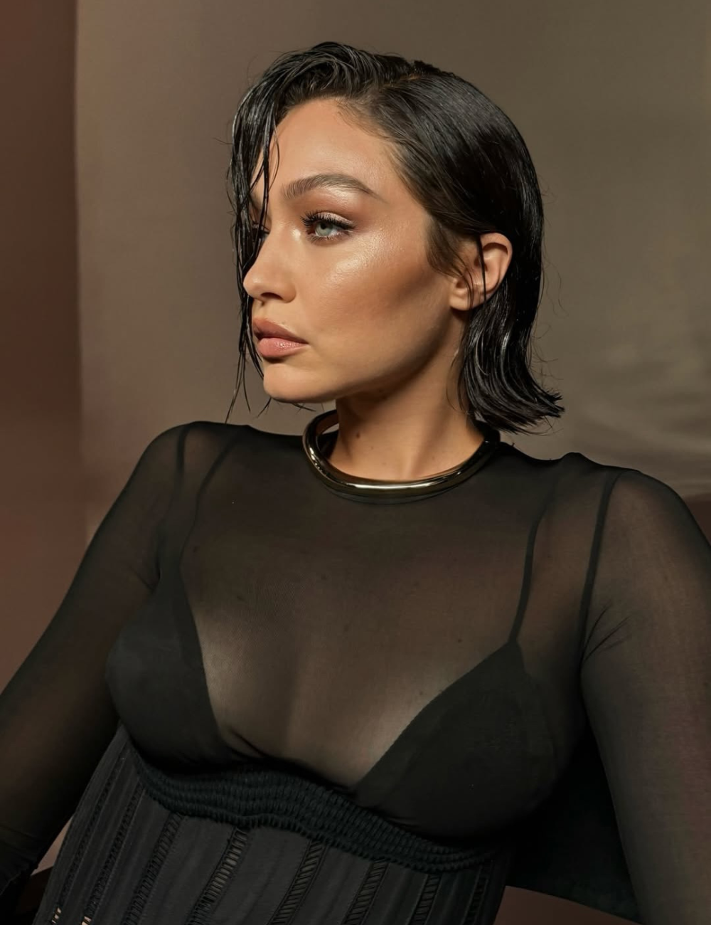 Modelo posa com blusa preta transparente de manga longa e top preto, cabelo molhado e maquiagem natural iluminada.