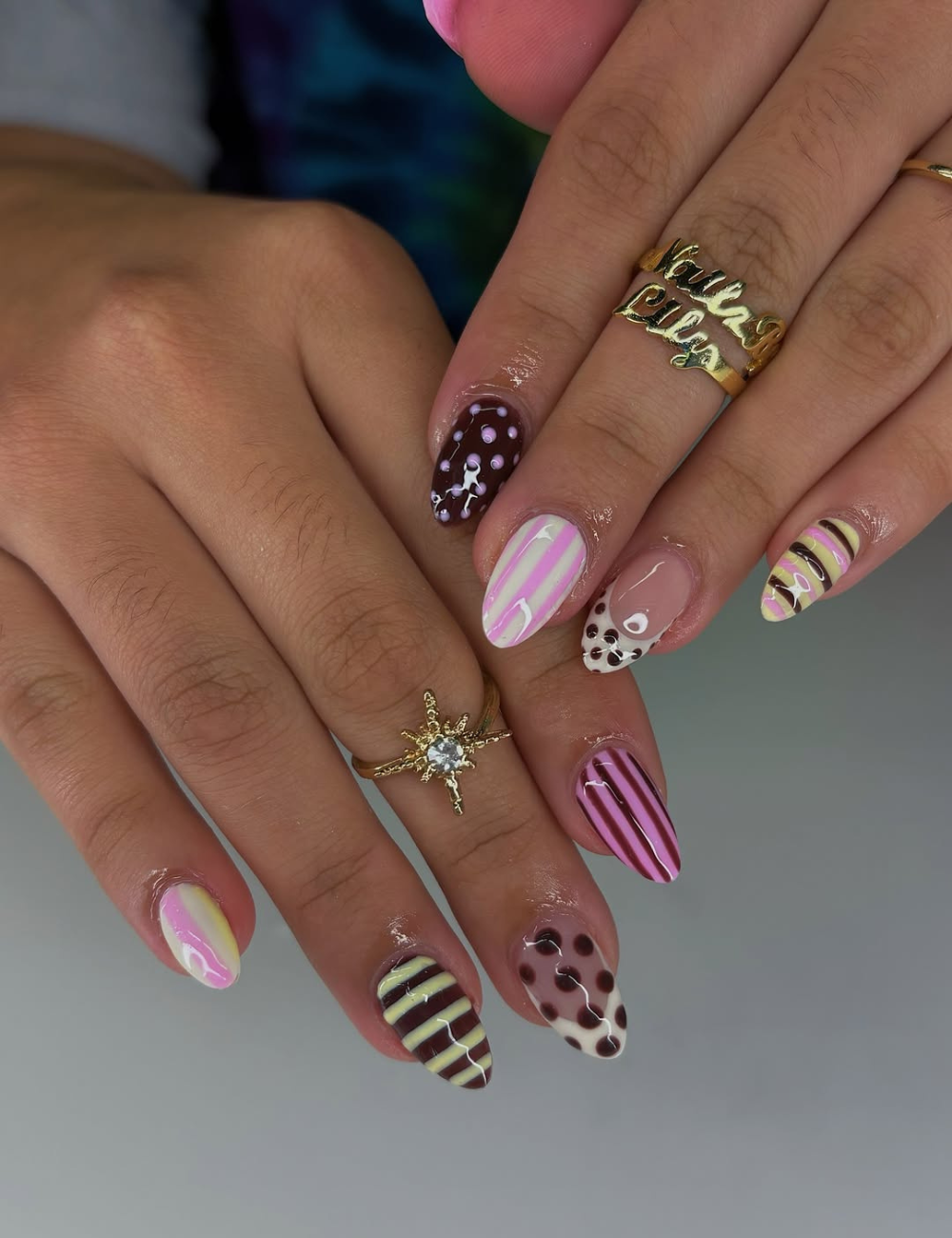 Unhas ovais decoradas com esmaltes em tons de rosa, marrom e amarelo, combinando listras e bolinhas.