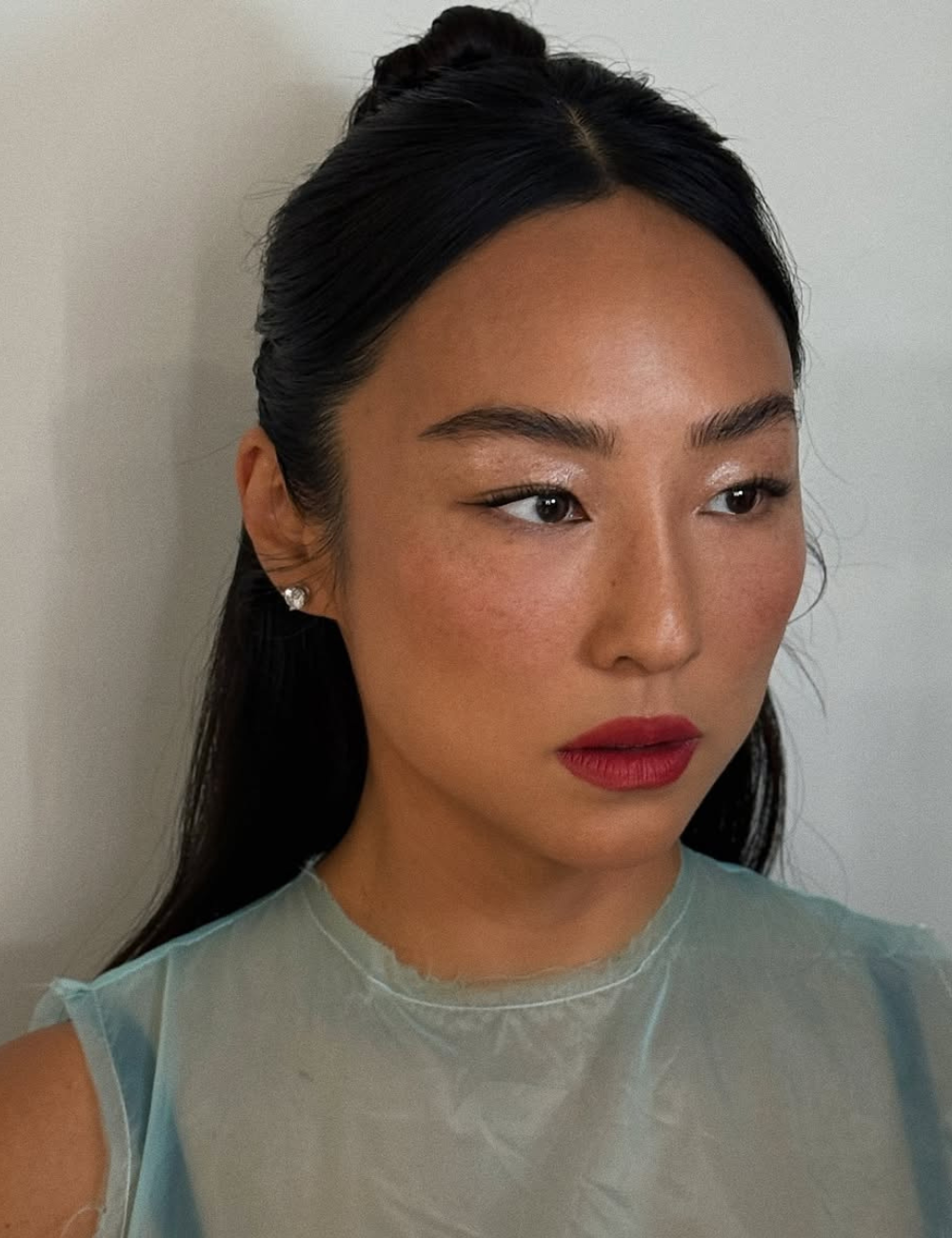 Modelo posa com maquiagem suave destacando pele natural e batom vermelho vibrante, usando blusa azul translúcida.