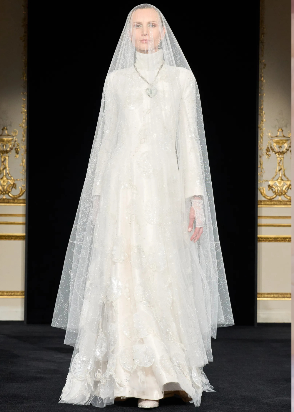 Modelo posa com vestido longo branco de alta-costura primavera 2026, gola alta, mangas longas, tecido bordado e véu