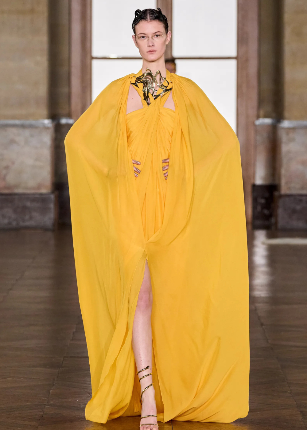 Modelo posa com vestido amarelo fluido de alta-costura primavera 2026, com capa ampla, fenda frontal e colar metálico