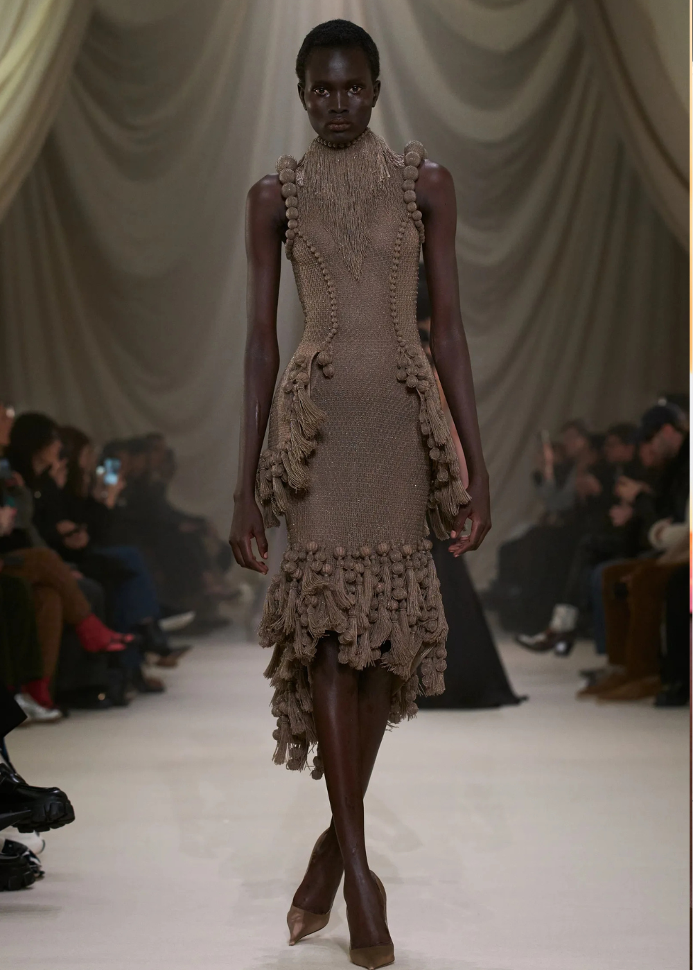 Modelo caminha com postura confiante usando vestido de tricô marrom com franjas e pompons, destacando tendências da