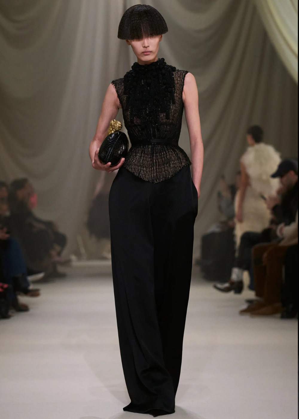 Modelo posa com blusa preta transparente estruturada e calça ampla, destacando tendências da temporada de alta-costura