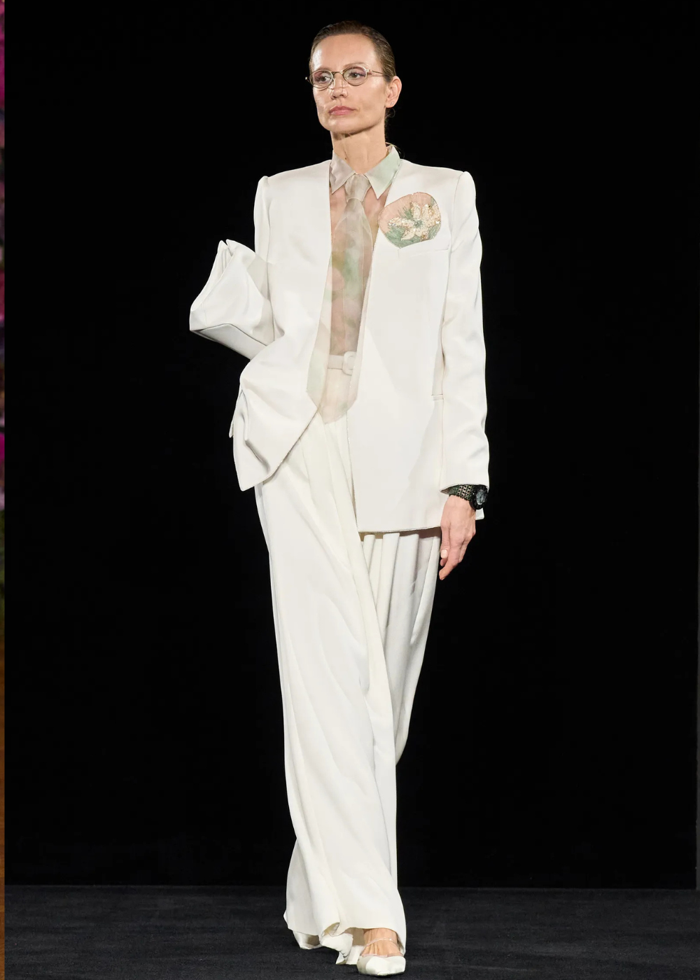 Modelo posa com terno branco oversized, gravata e camisa translúcida em tons pastel, destacando elegância e sofisticação nas