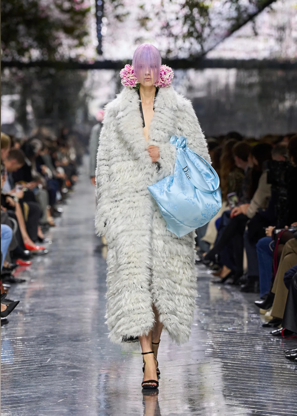 Modelo caminha com postura firme e expressão séria, vestindo casaco volumoso de pelúcia branca, bolsa azul celeste de cetim