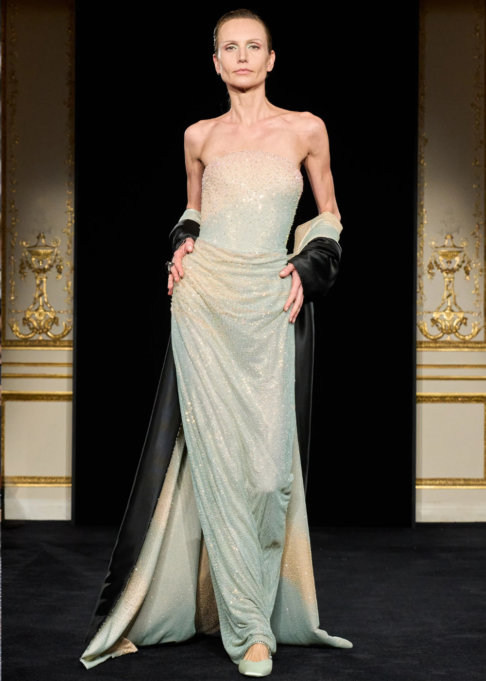 Modelo posa com vestido tomara que caia em tecido brilhante azul claro esverdeado e manto preto, expressando sofisticação