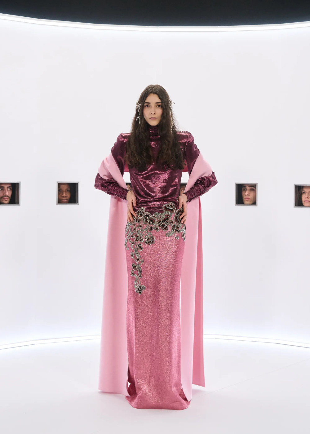 Modelo posa com vestido longo rosa cintilante e blusa vinho metálica, destacando bordados florais e capa rosa, expressando